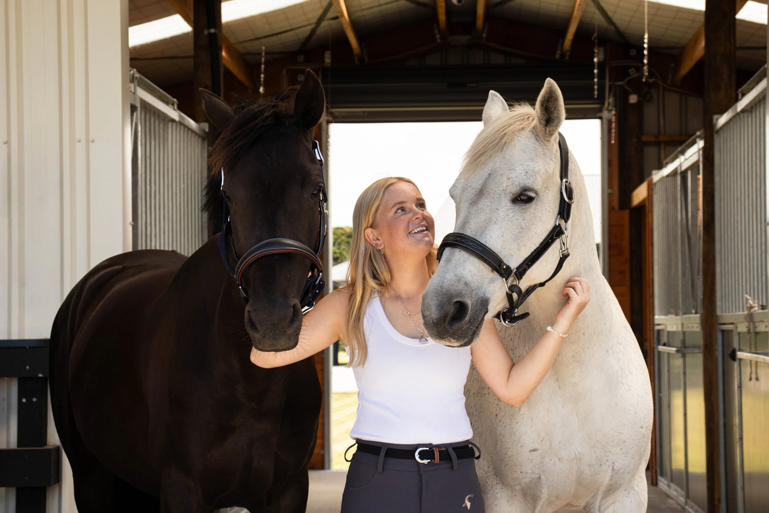 A woman smiling with two horses, one black and one white, inside a horse stable.