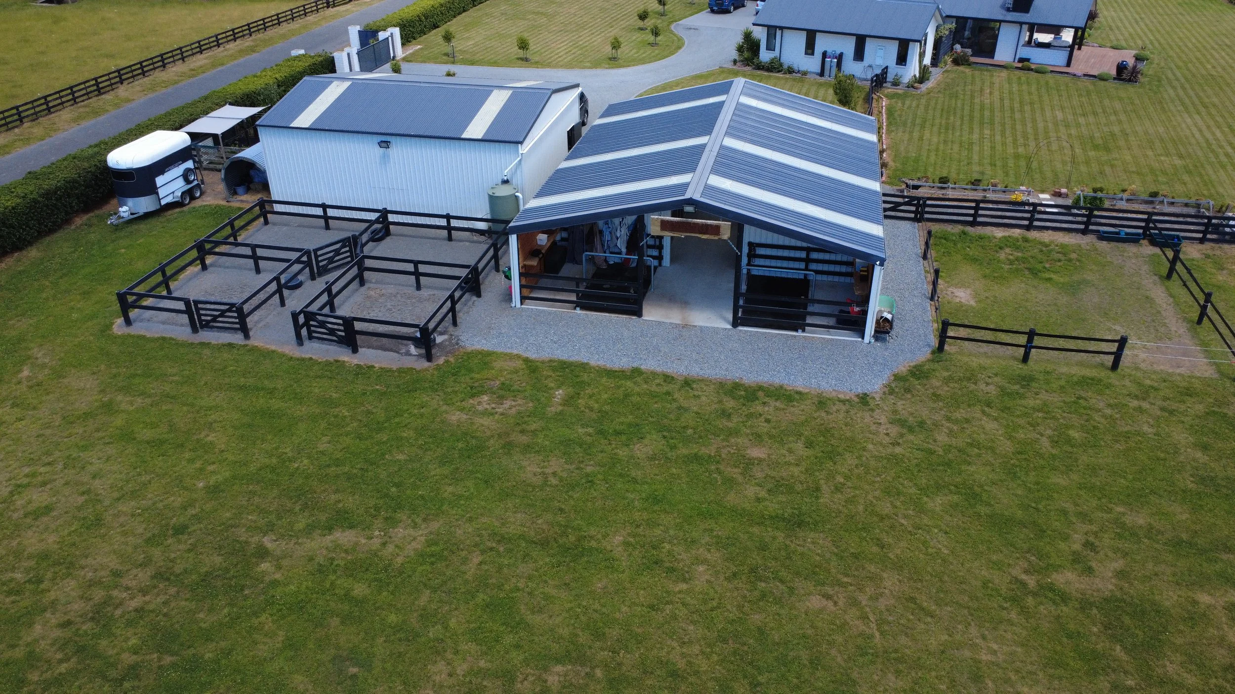 Aerial view of a farmyard showing a covered area with fencing, a white metal shed, a second building with a blue roof, a trailer, and a grassy yard surrounding the structures.