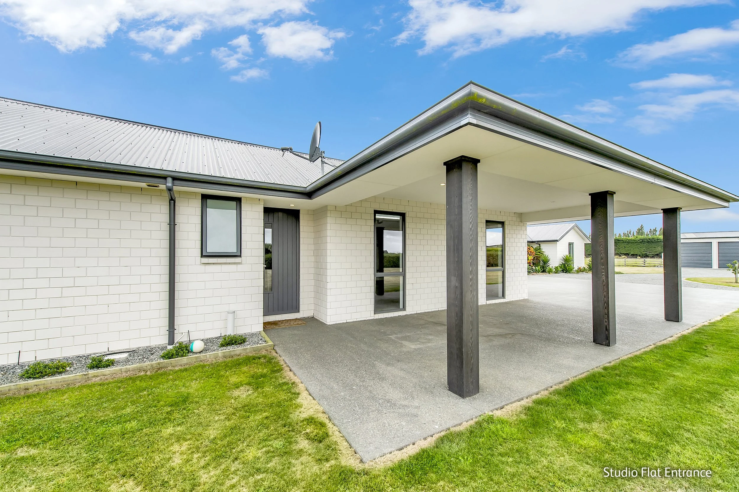 Front view of a modern house with a covered porch supported by black wooden pillars, white brick exterior, metal roof, and a small garden bed, under a partly cloudy sky