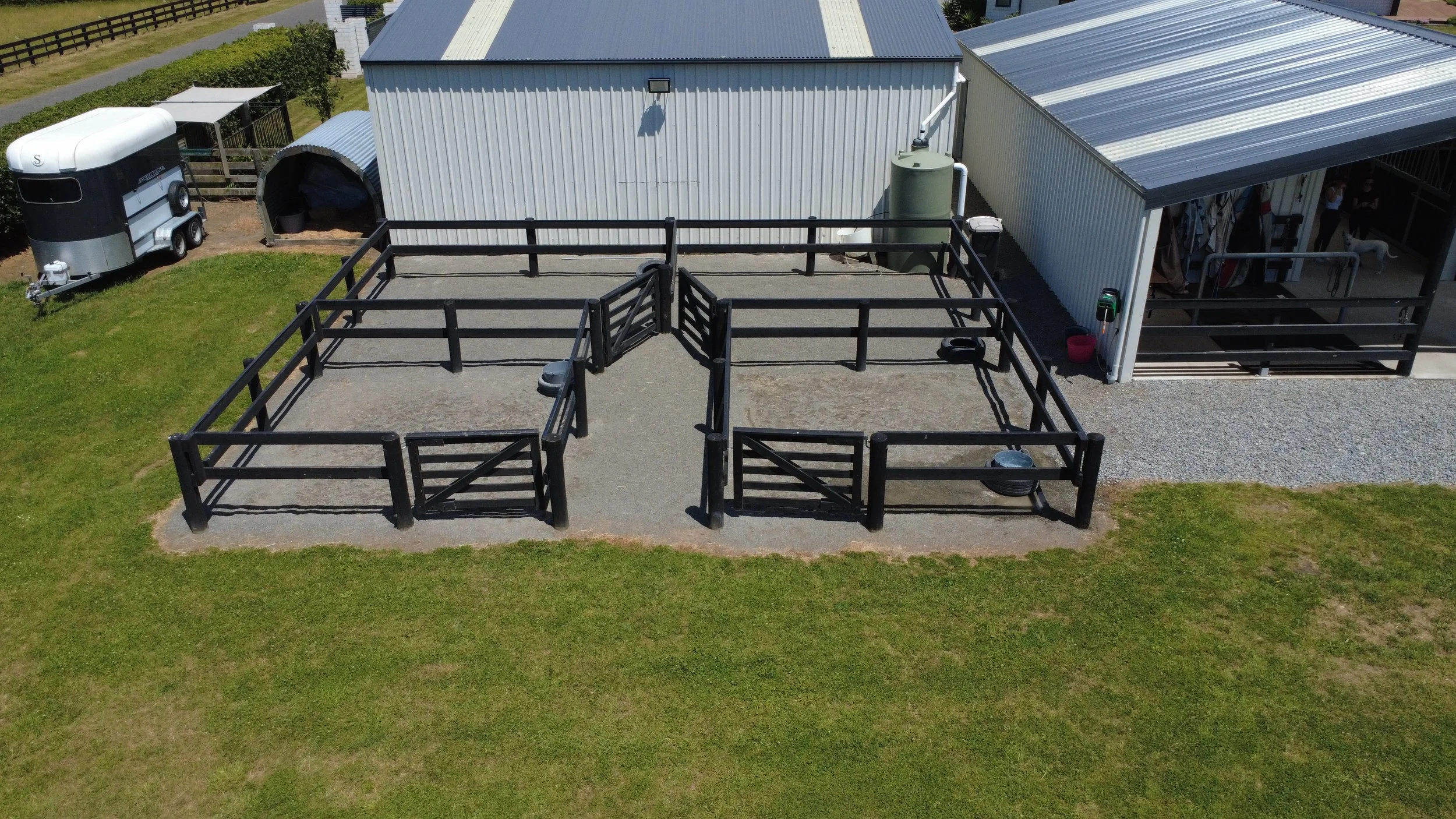 Aerial view of an outdoor horse riding arena with black fencing, adjacent to a white barn and covered area, on a grassy field with some gravel patches.