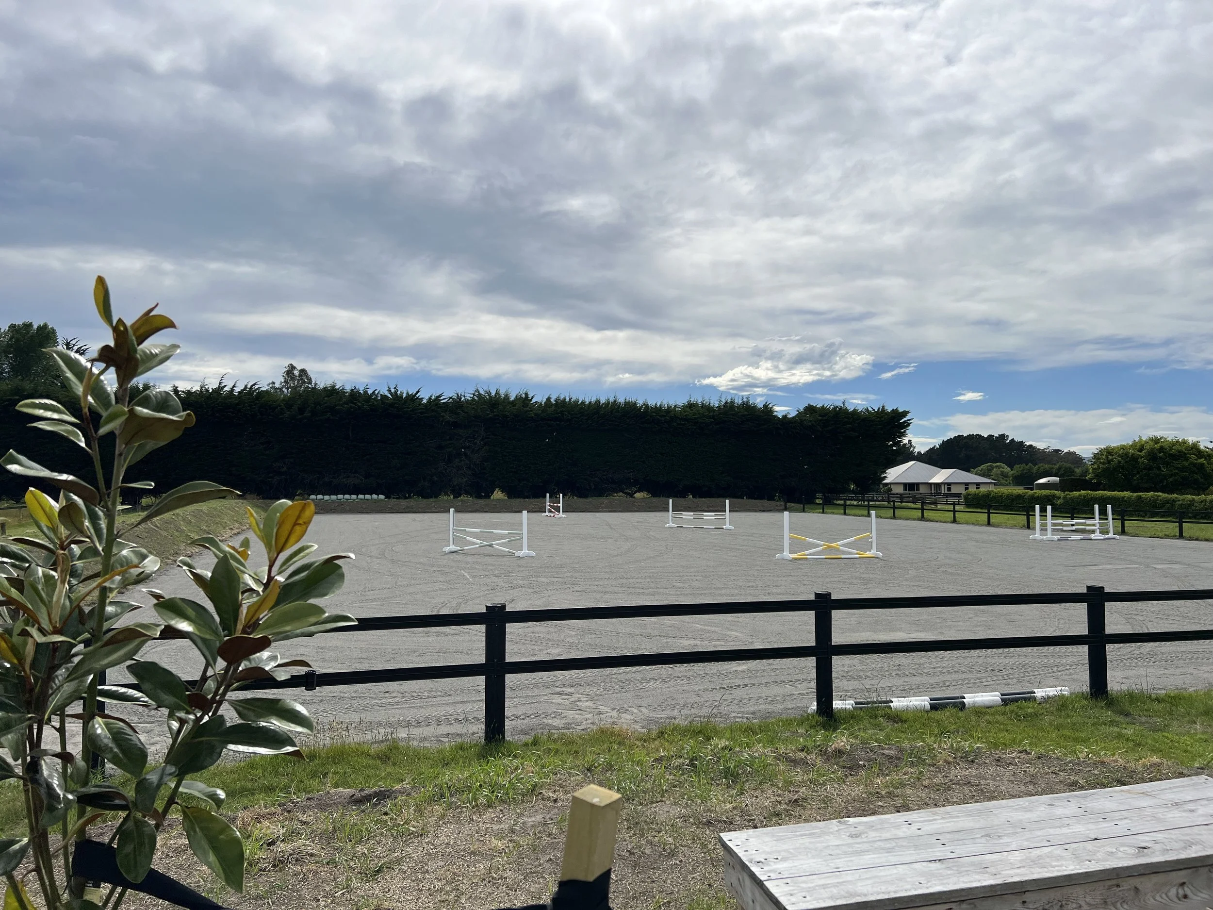 An outdoor equestrian show jumping arena with several white jumps, surrounded by a black fence, under a partly cloudy sky during daytime.