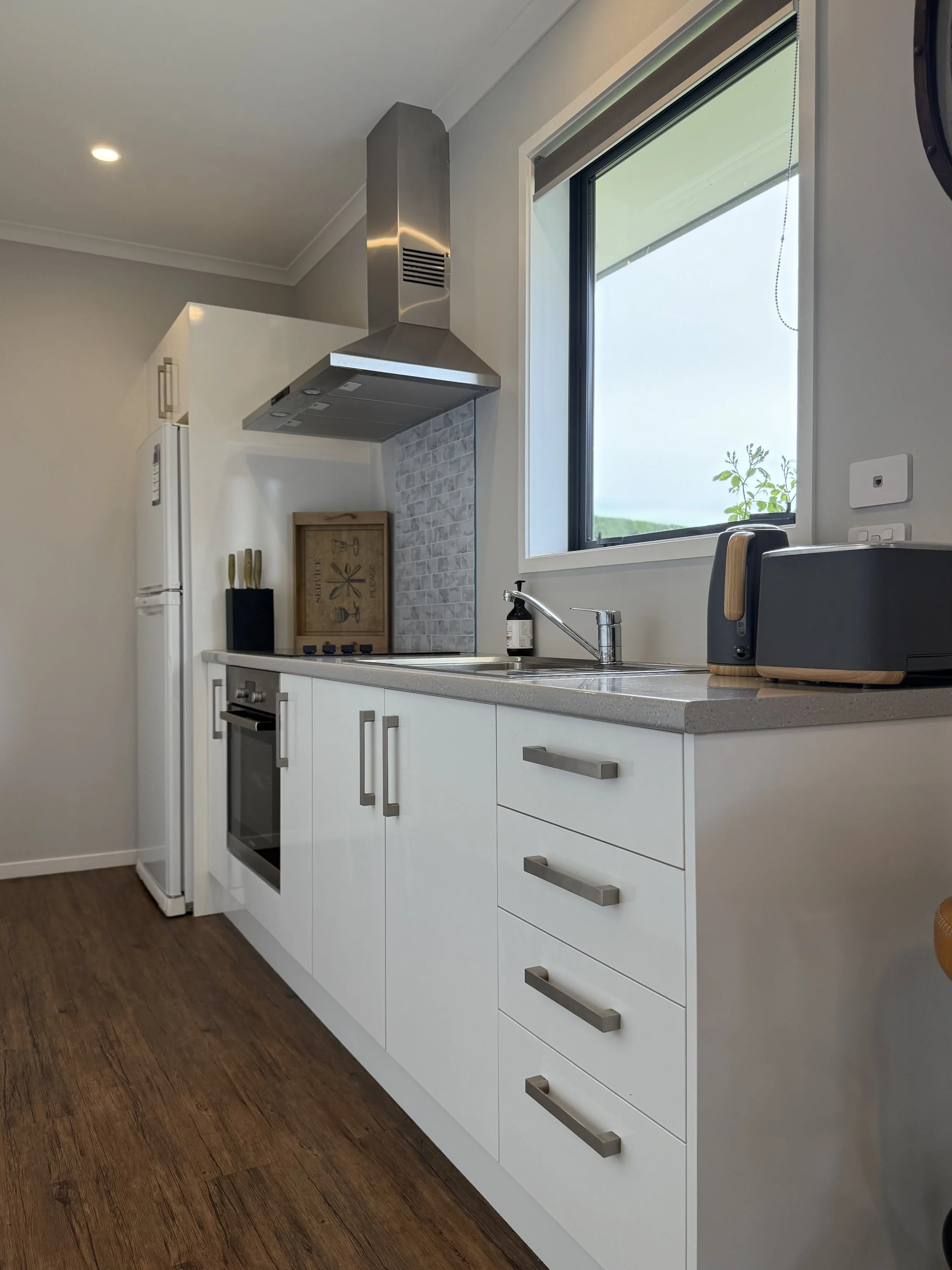 Modern kitchen with white cabinets, stainless steel range hood, window, and wooden flooring.