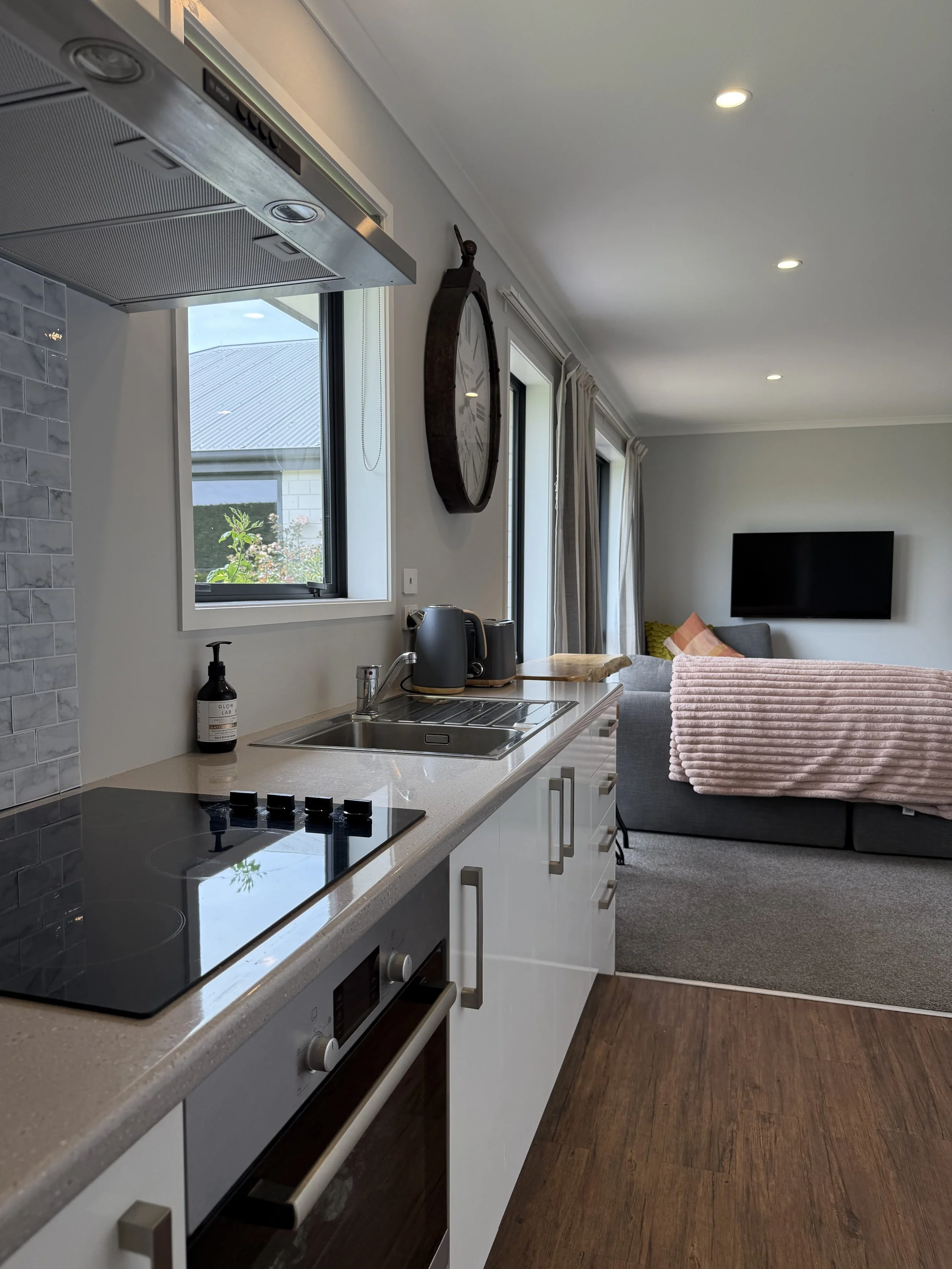 Open kitchen with white cabinets, black stovetop, and a microwave oven, with a living room area visible in the background, including a gray couch, cushioned blanket, and flat-screen TV on the wall.