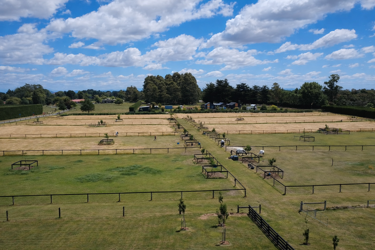 Horse riding arena with fenced sections and people riding horses, set in a lush green landscape under a partly cloudy sky.