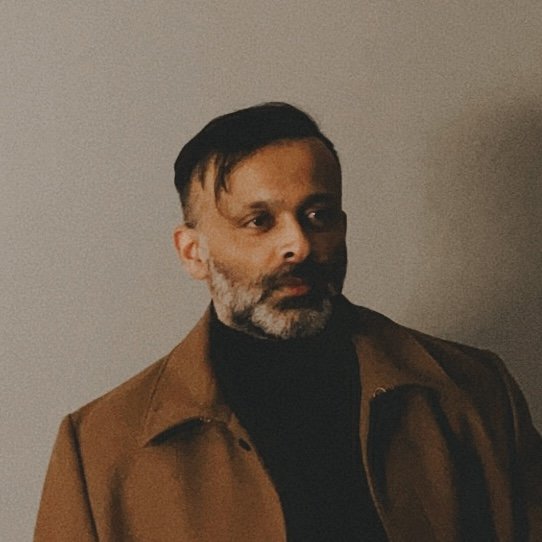 A man with dark hair and a beard, wearing a brown coat over a black turtleneck, standing against a neutral background.
