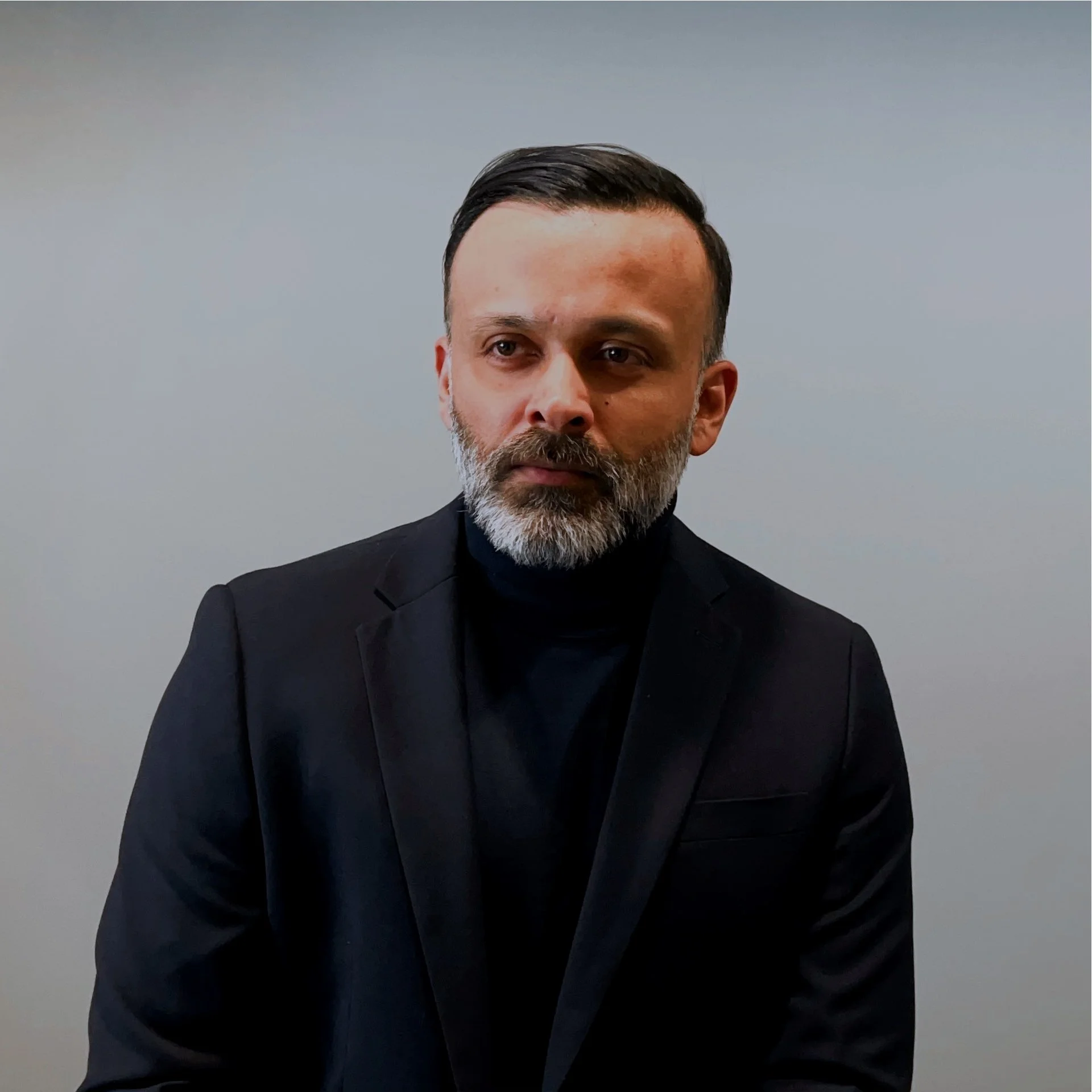 Professional portrait of a middle-aged man with dark hair and a gray beard, wearing a black suit and black shirt, against a plain light background.