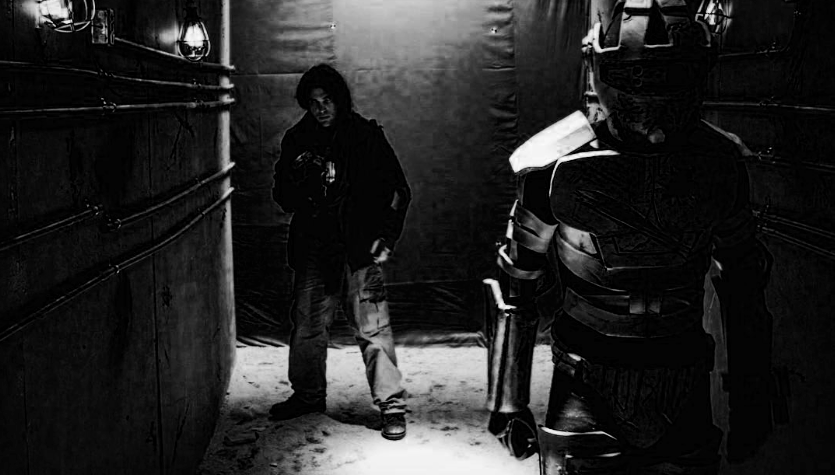 A robot in a futuristic or armored costume, facing another person in a dark corridor or room with minimal lighting, walls lined with pipes or wires.