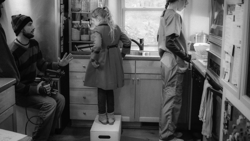 A film director with a camera recording a young actress in a kitchen with a woman preparing food.