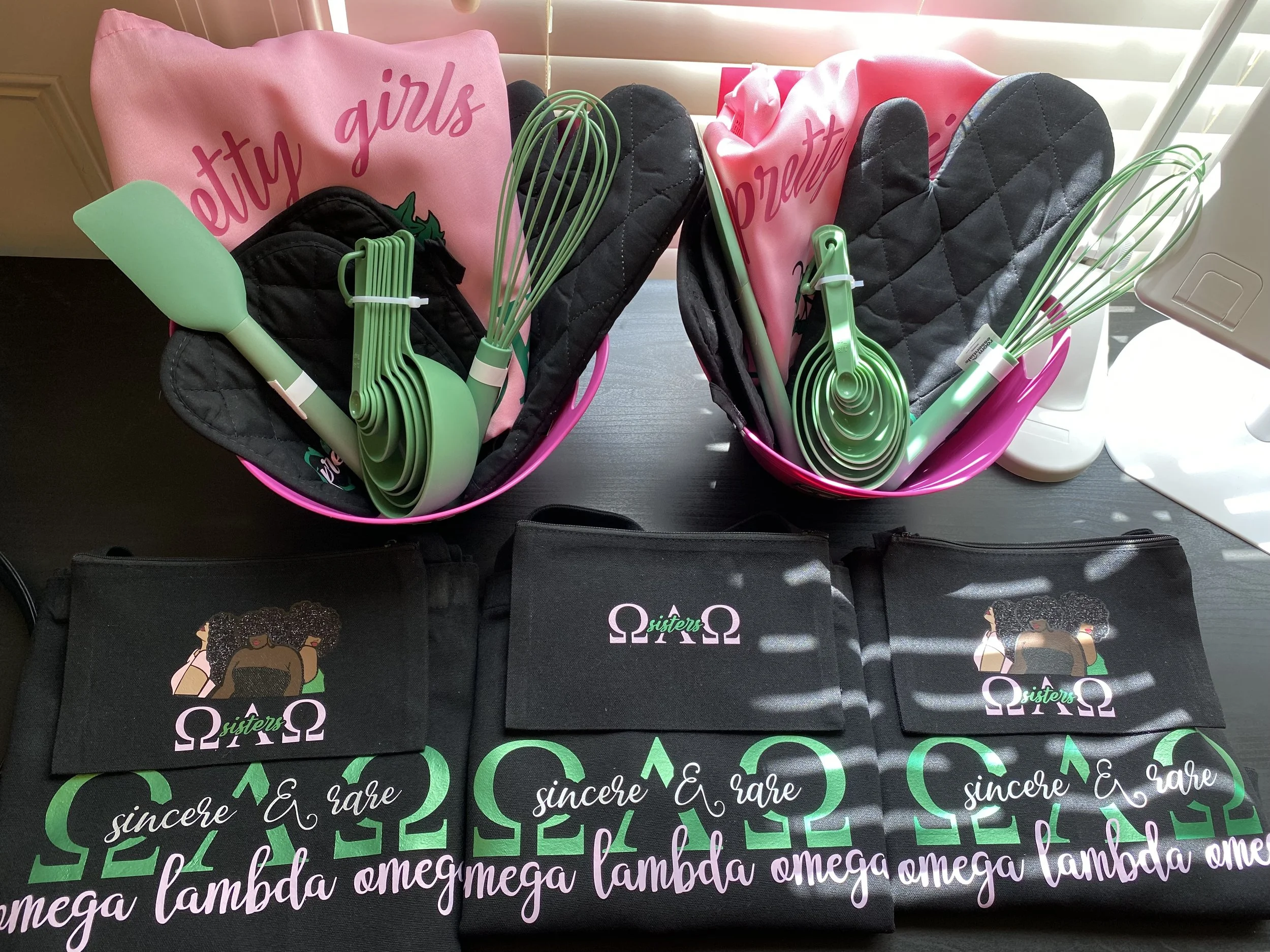 Two pink bowls contain green silicone kitchen tools, including spatulas and whisks, along with two pink cloths with the words 'pretty girls' printed on them. On the table in front, there are three black fabric pouches with a design of diverse women a