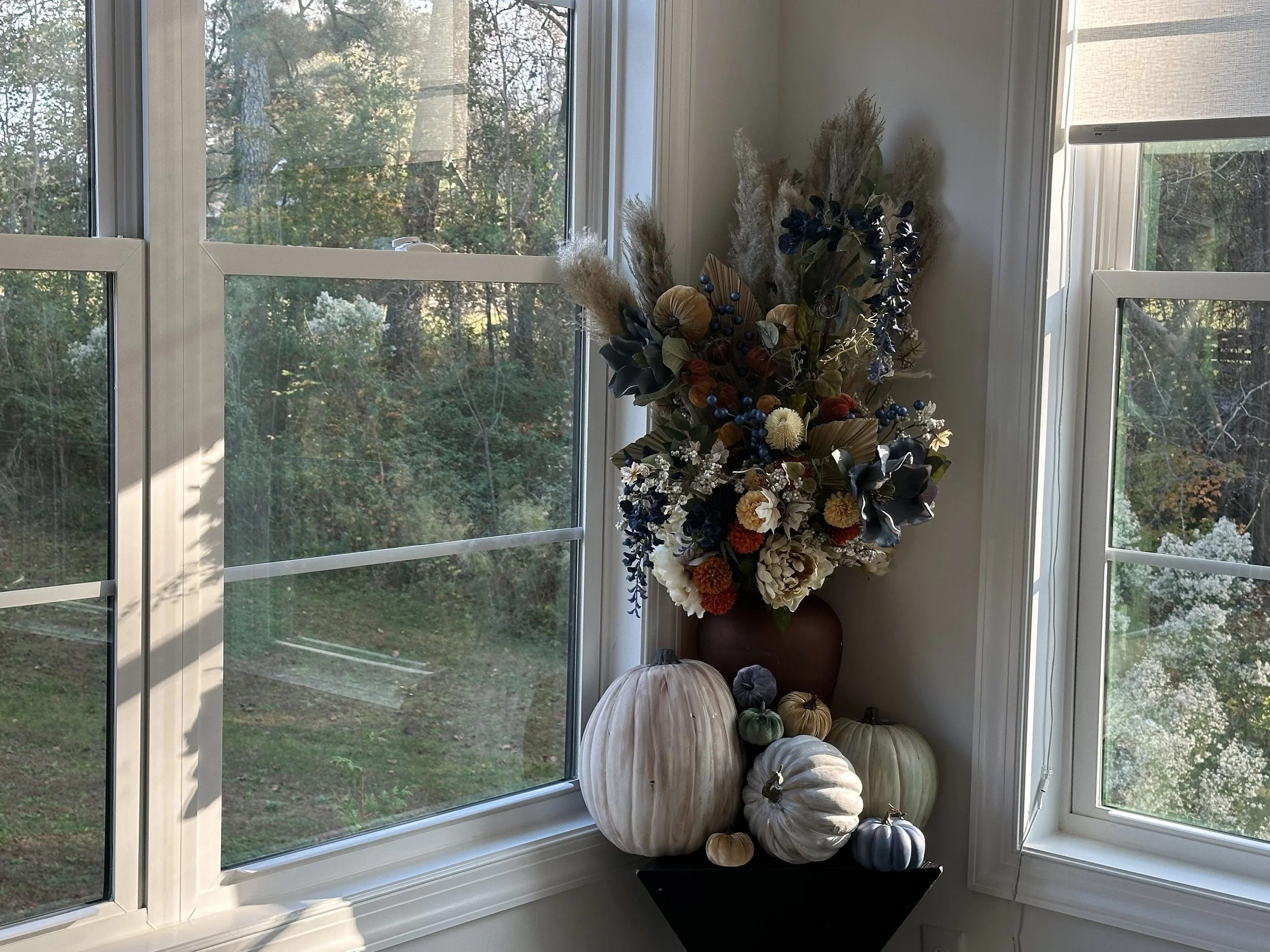 Decorative fall arrangement with dried flowers and various colored pumpkins on a black stand near a corner with large window.