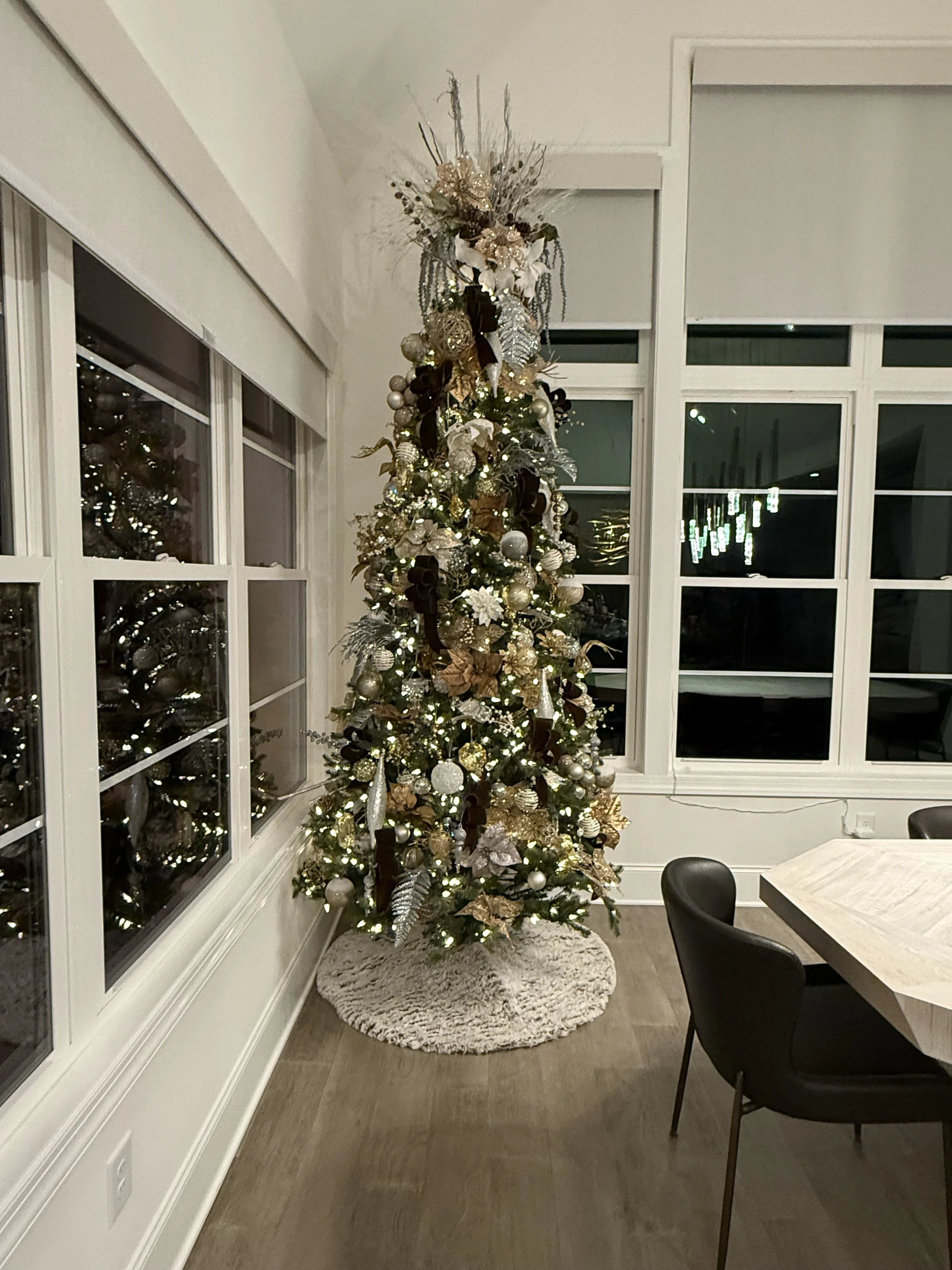 Decorated Christmas tree near windows in a modern living room, with ornaments in gold, silver, and white, and a fluffy white tree skirt.