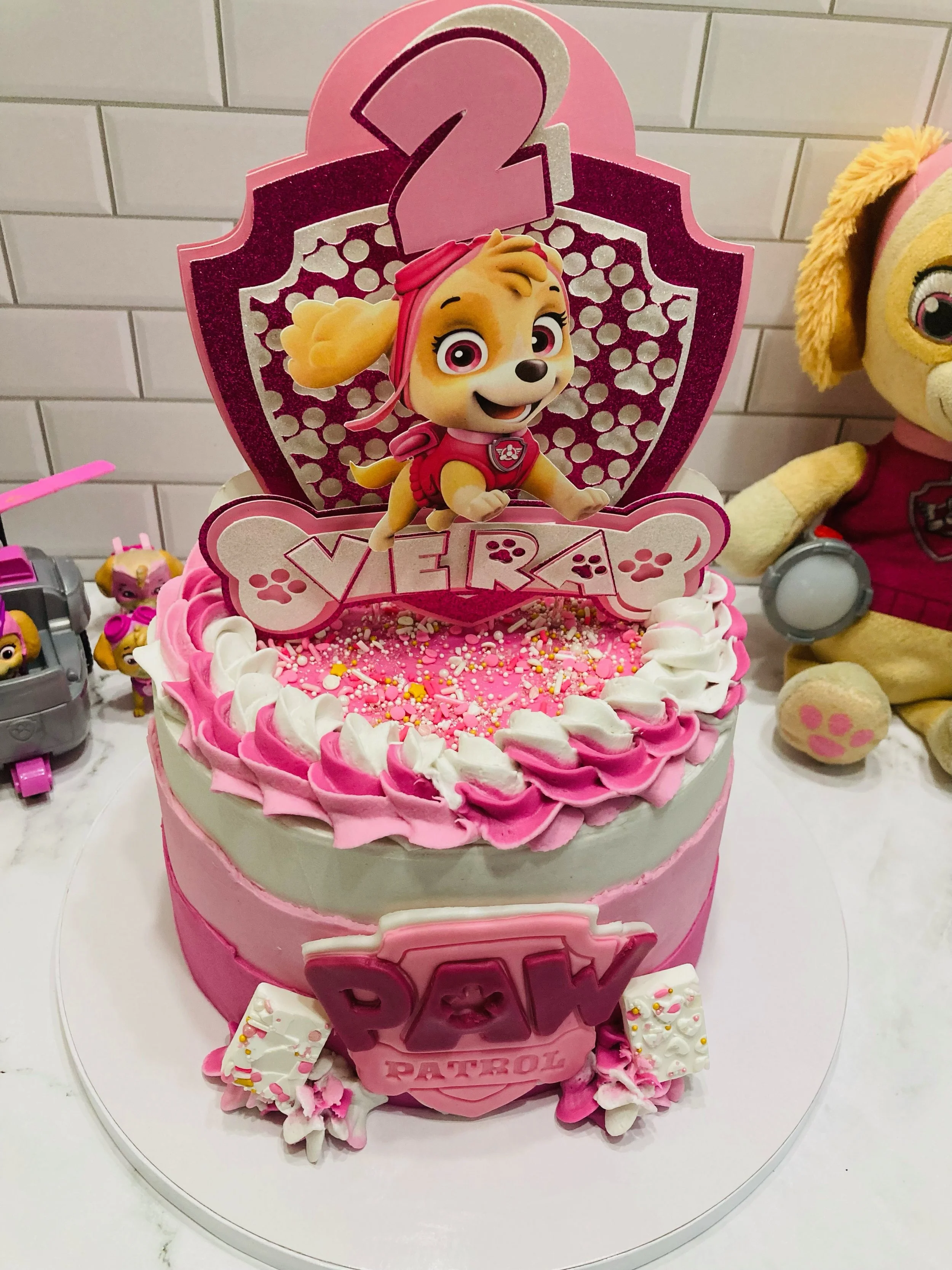 Pink birthday cake decorated with white and pink icing, sprinkles, and a topper featuring Skye, a character from TV show PAW Patrol, with a large pink number 2.