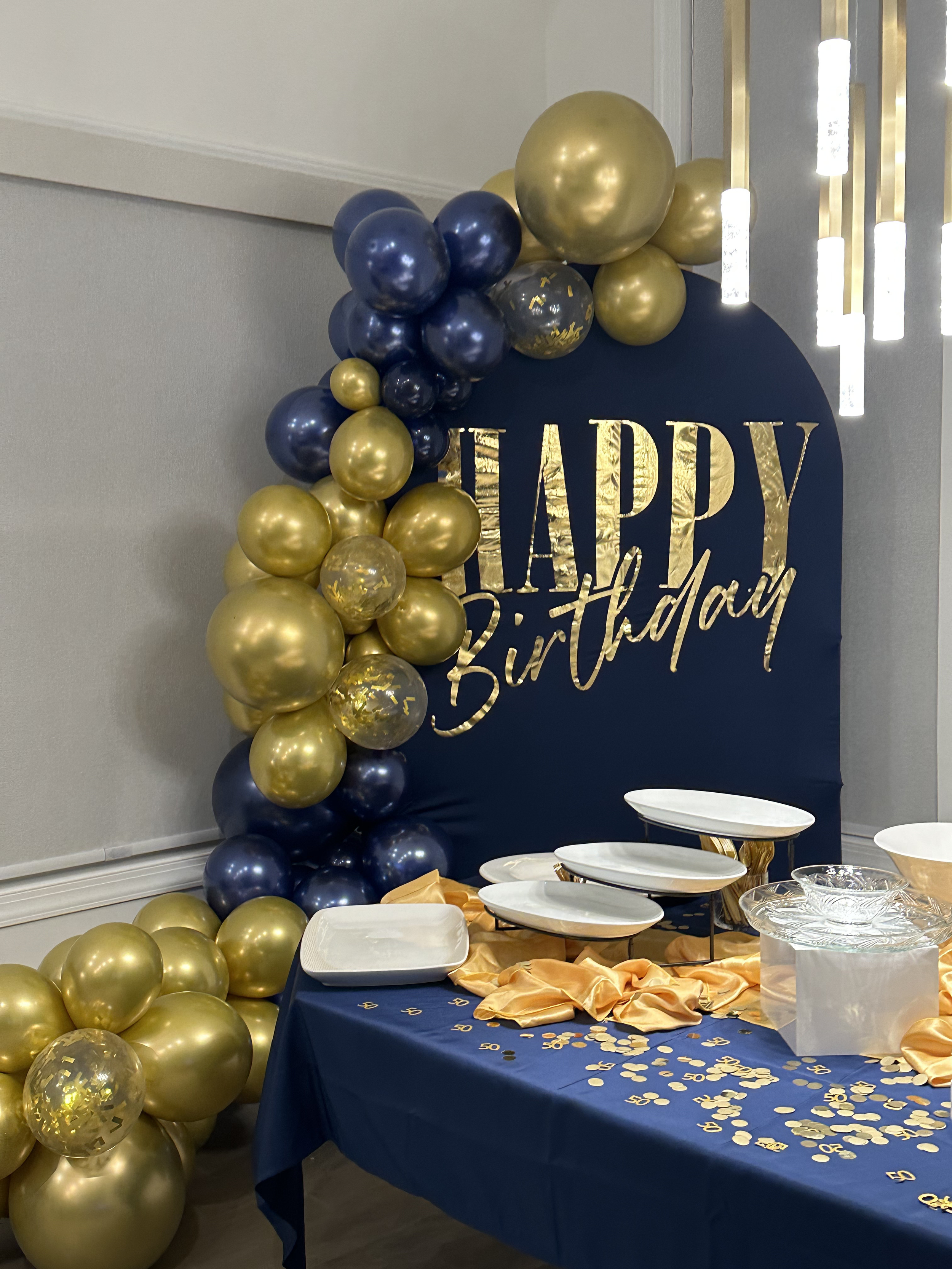 Blue and gold birthday decoration with balloons, a navy blue backdrop with gold lettering that reads "HAPPY Birthday", and a table with plates, gold confetti, and a gold satin cloth.