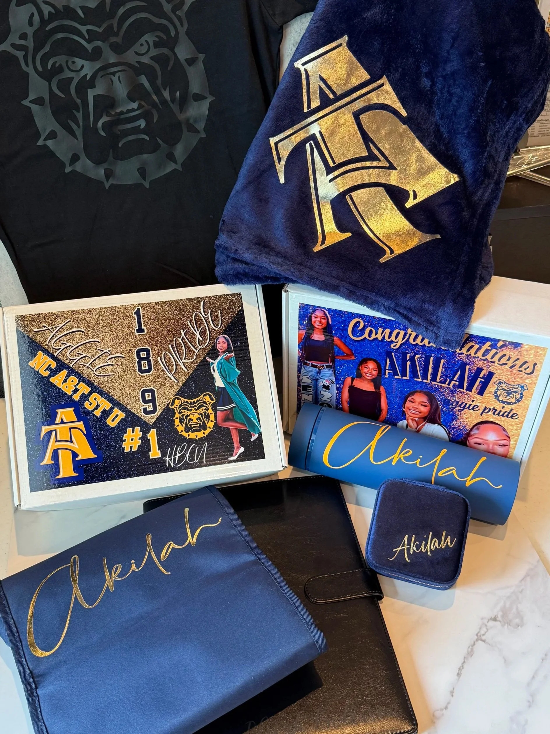 A collection of items celebrating a graduation, including a black shirt with a bulldog logo, a blue towel with a gold letter 'A', a personalized box with photos and the name 'Akhah', a blue pen holder with 'Akhah', a black planner, and a blue cloth w