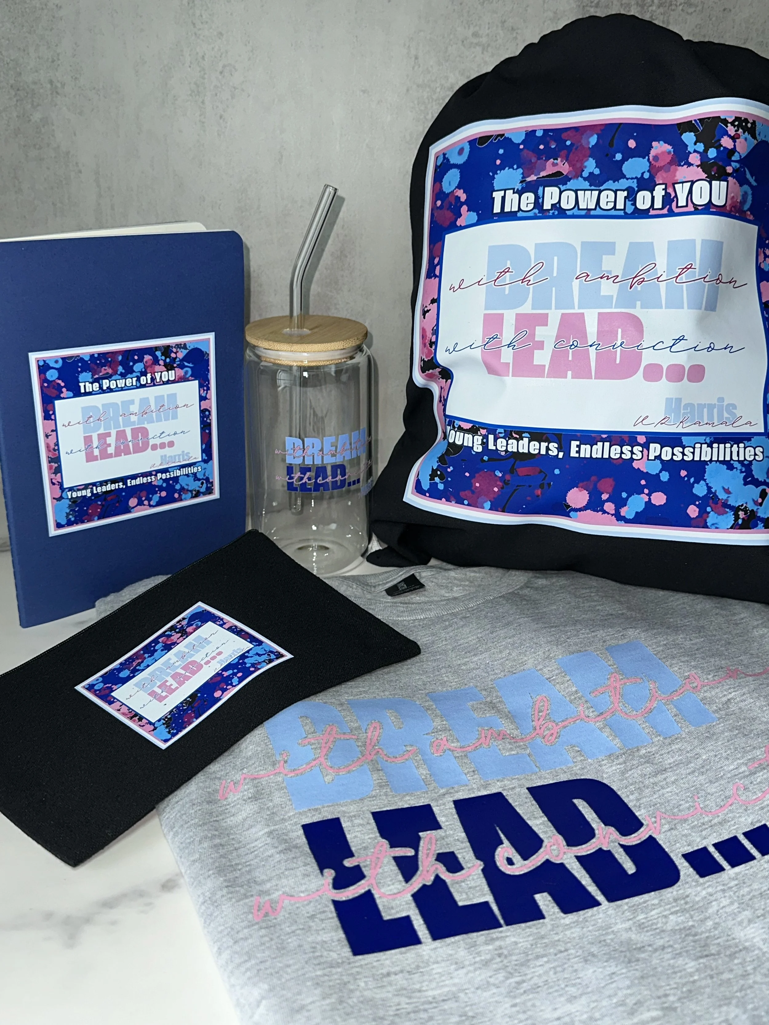 Collection of items including a backpack, a tumbler with a straw, a notebook, a cloth with a printed message, and a folder. All items feature the same colorful design with blue, pink, and purple splatters and the text 'The Power of YOU, Dream BIG, Le