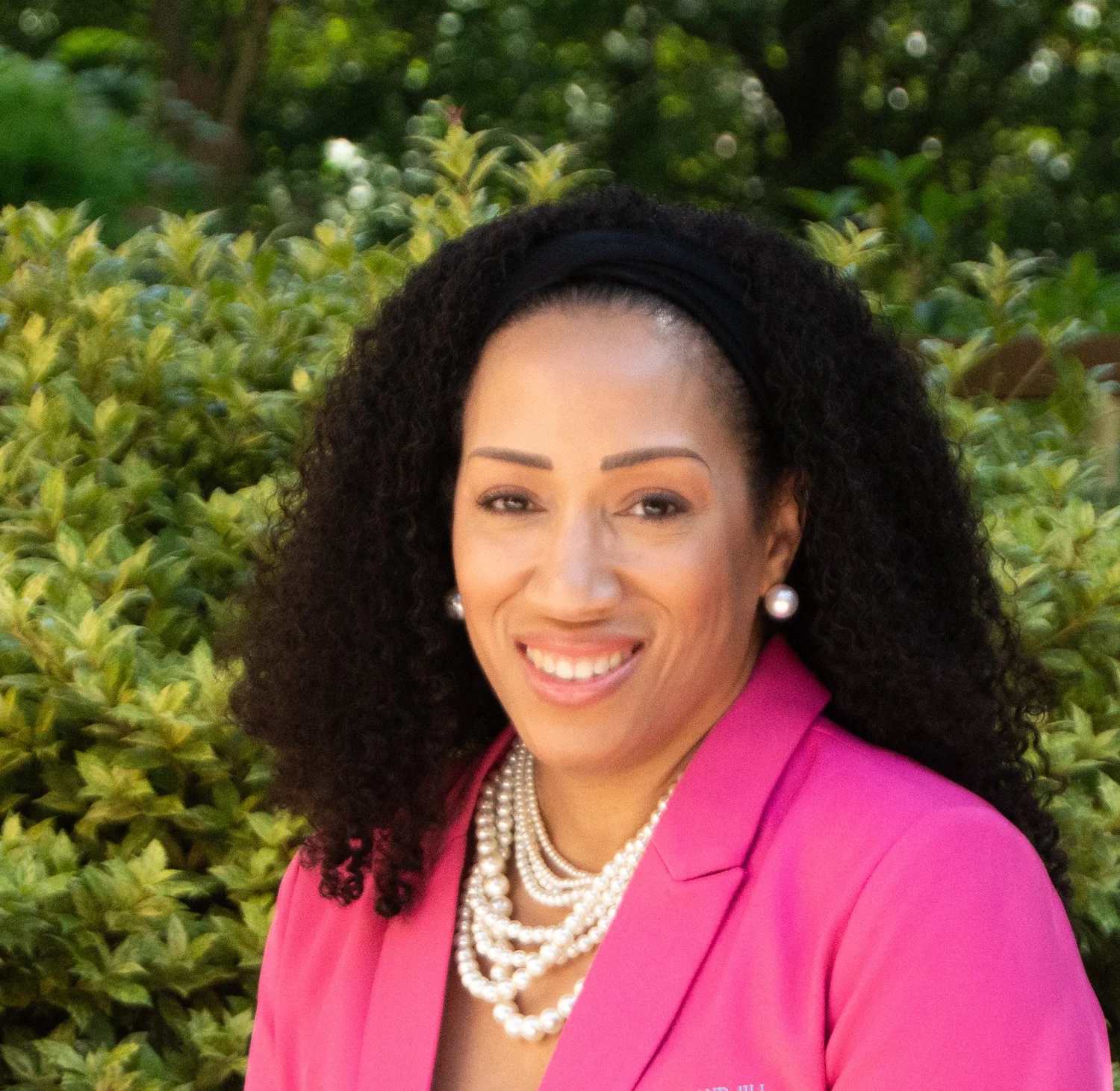 A woman with curly black hair, wearing a pink blazer, pearl necklace, pearl earrings, and a black headband, smiling outdoors with green bushes in the background.