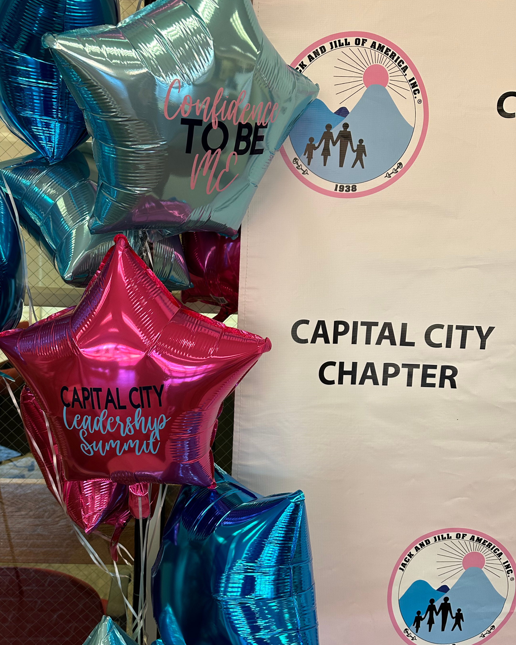 Colorful star-shaped balloons with text and event logos in front of a banner that reads 'Capital City Chapter.' The balloons have messages like 'Confidence to Be Me' and 'Leadership Summit.'