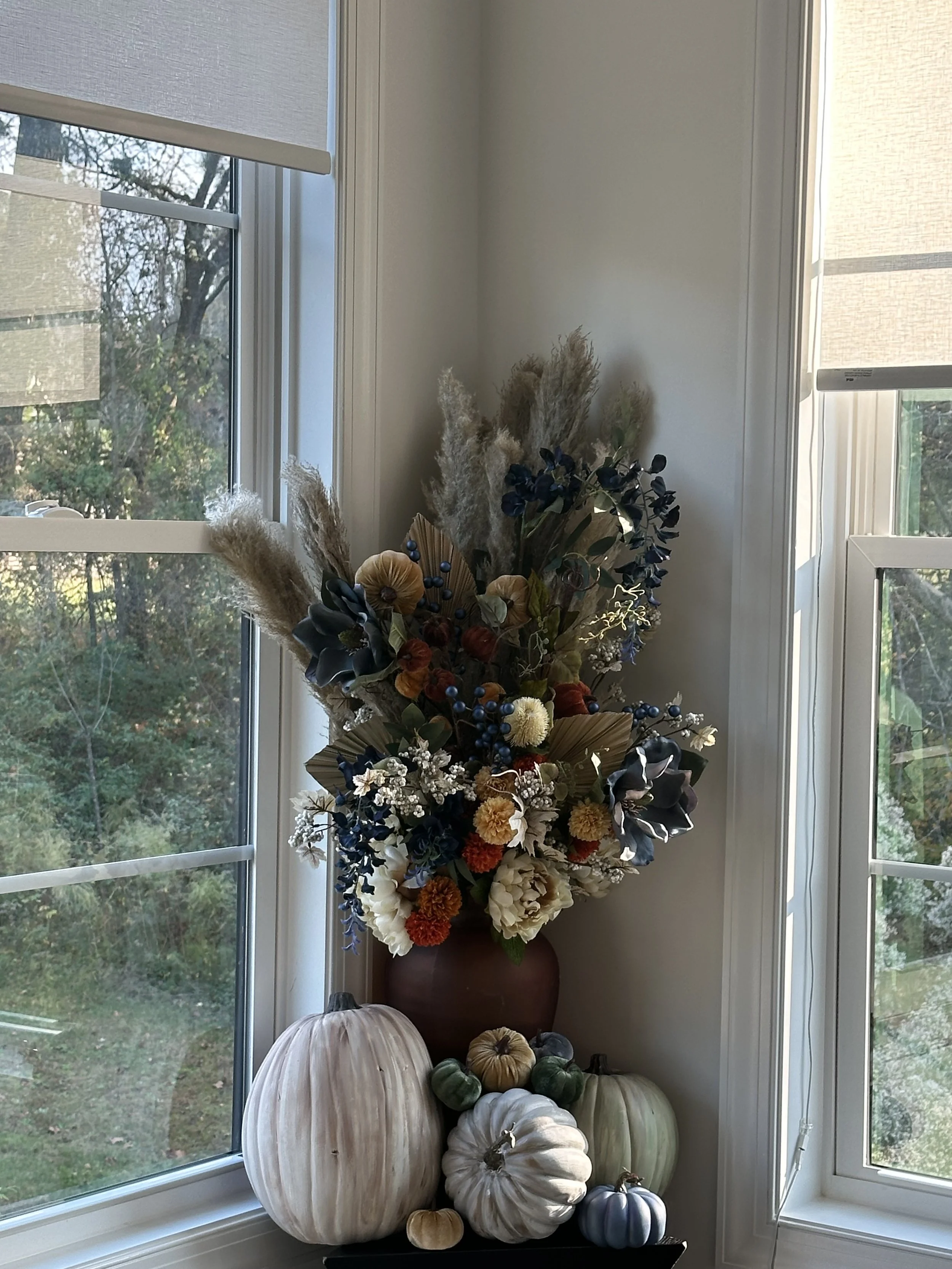 A large floral arrangement in a brown vase with dried and artificial flowers, surrounded by white, green, orange, and blue pumpkins, placed on a black surface by a corner window.