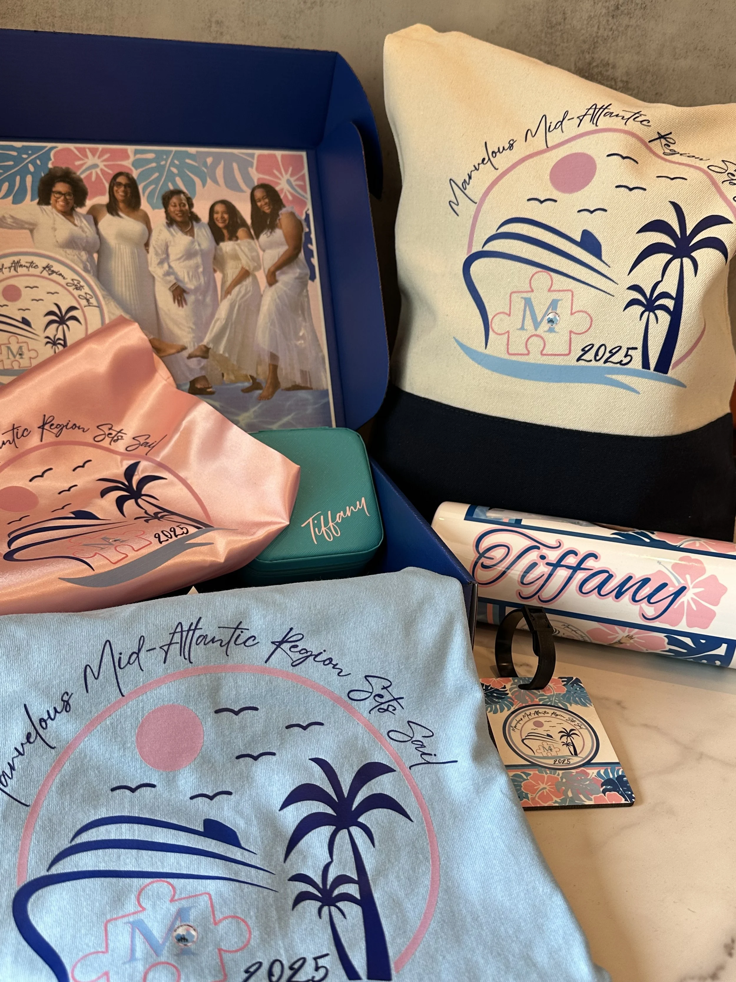 Items for the Mid-Atlantic Region New Year event in 2025, including a photo of six women dressed in white, various branded textiles with a tropical sunset and palm trees, a small tin box, and a keychain.