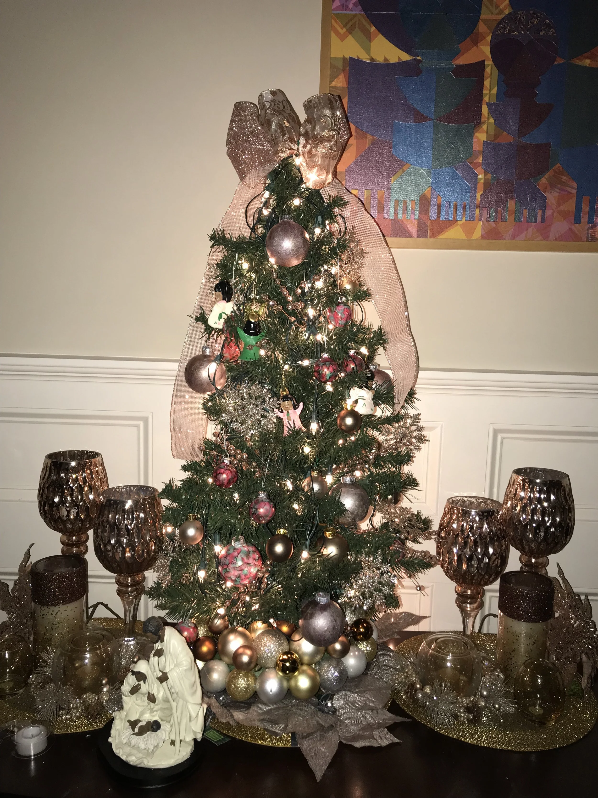Decorated Christmas tree with ornaments, lights, and a large pink bow on top, surrounded by candles and decorative items on a table.
