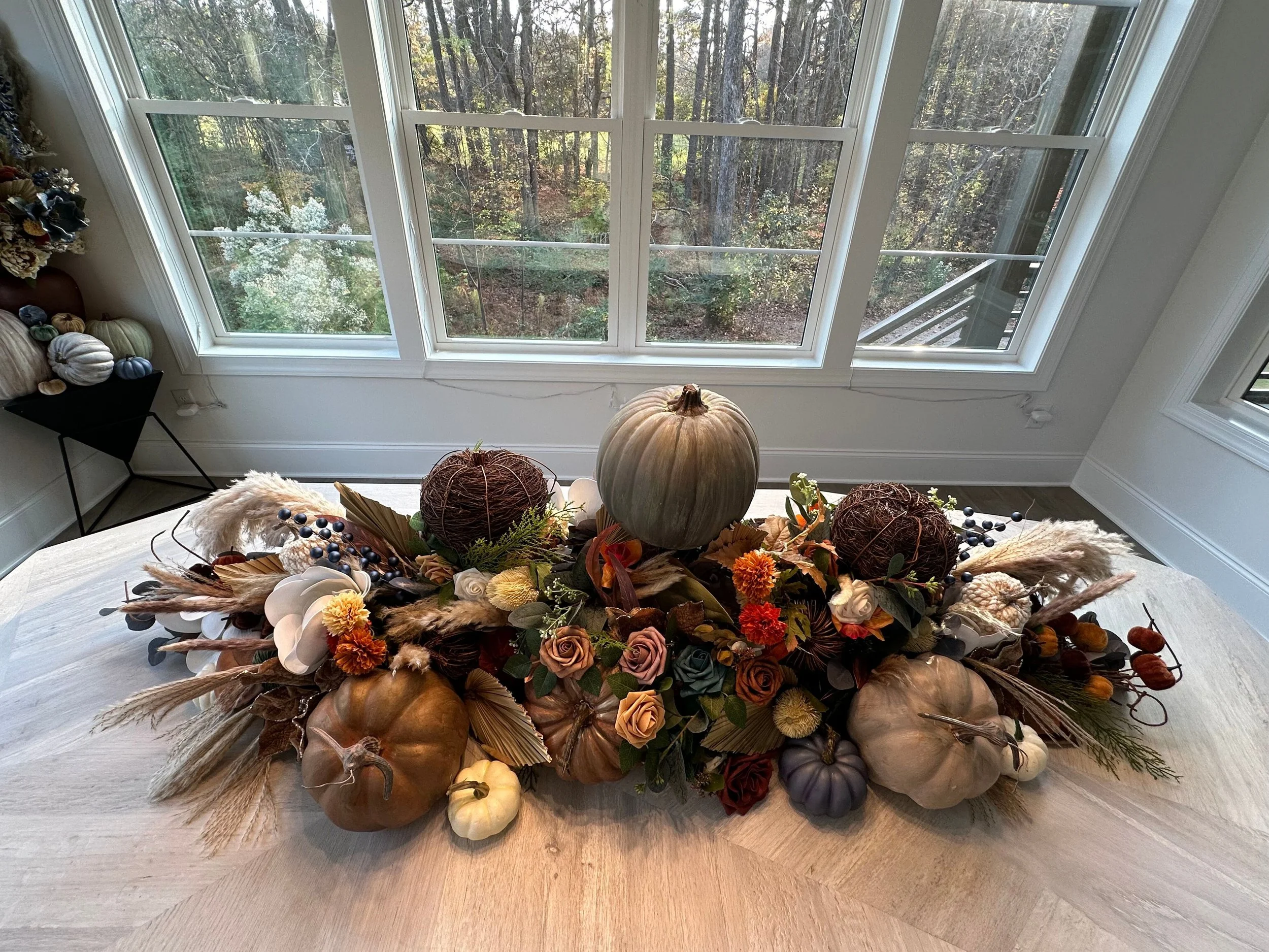 Autumn floral and pumpkin centerpiece on a light wood table, with large windows showing trees outside.