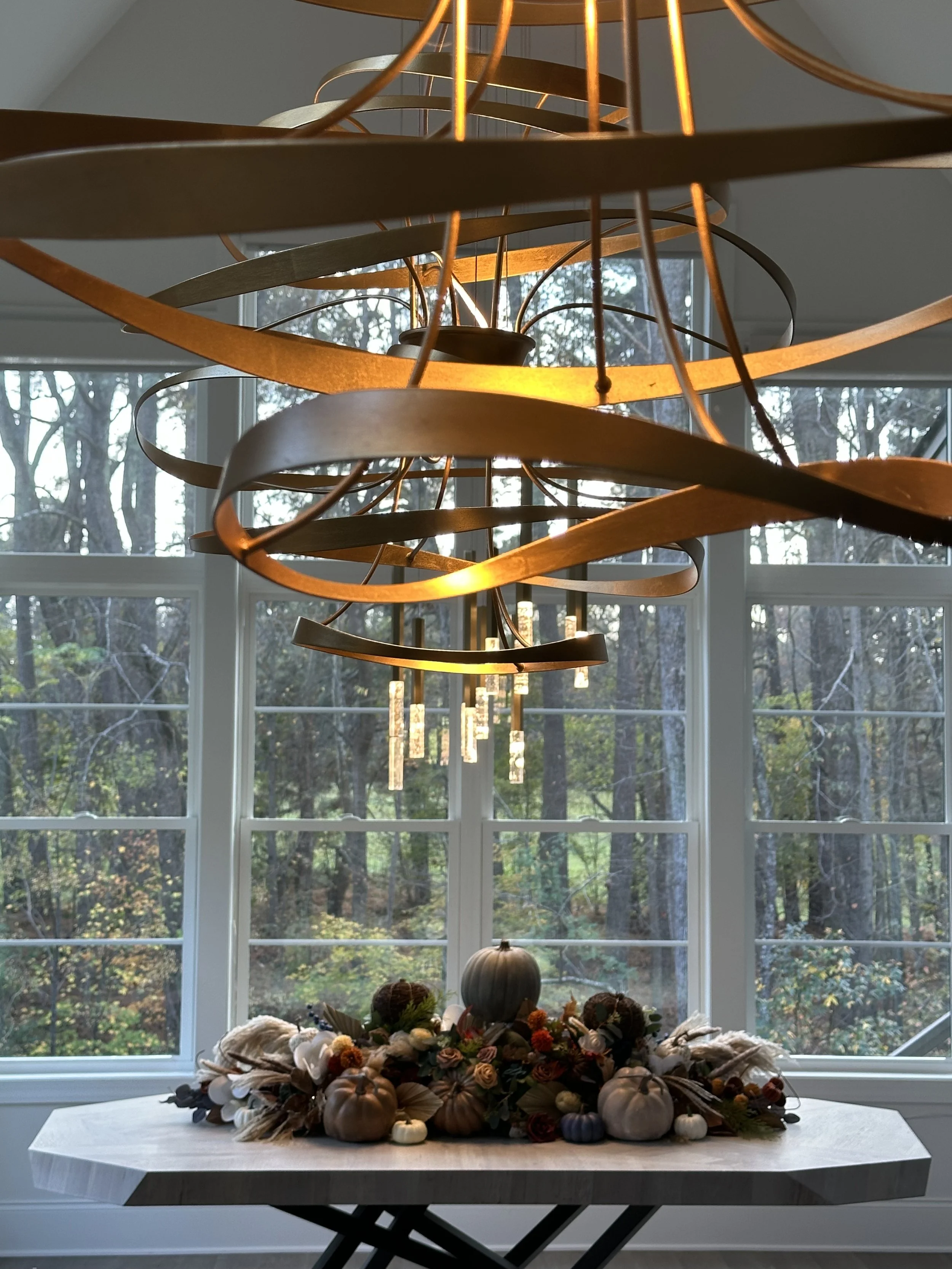 Interior view of a modern chandelier with gold-colored rings hanging above a decorated pumpkin centerpiece on a table, with large windows showing trees outside.