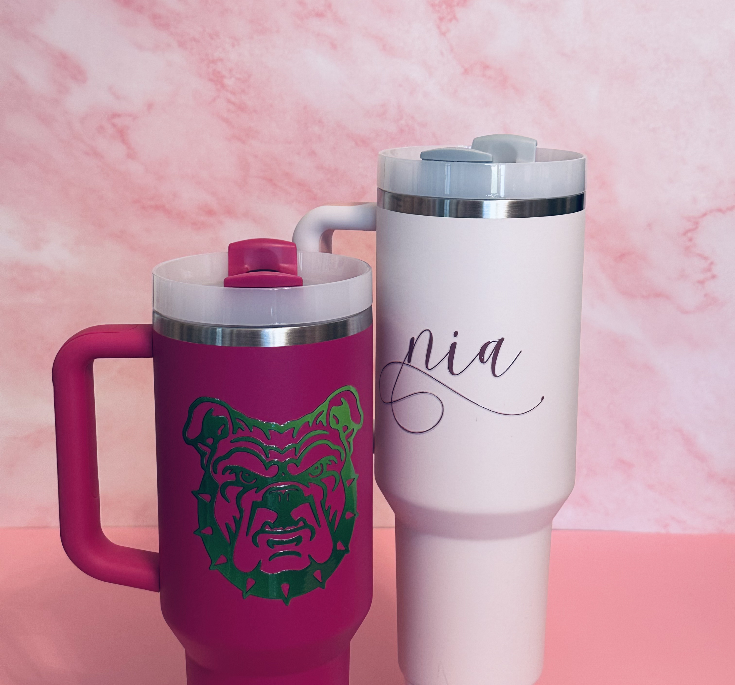 Pink and white insulated travel mugs with custom designs; the pink mug features a bulldog face, and the white mug has the handwritten name 'Nia'; both are set against a pink marbled background.