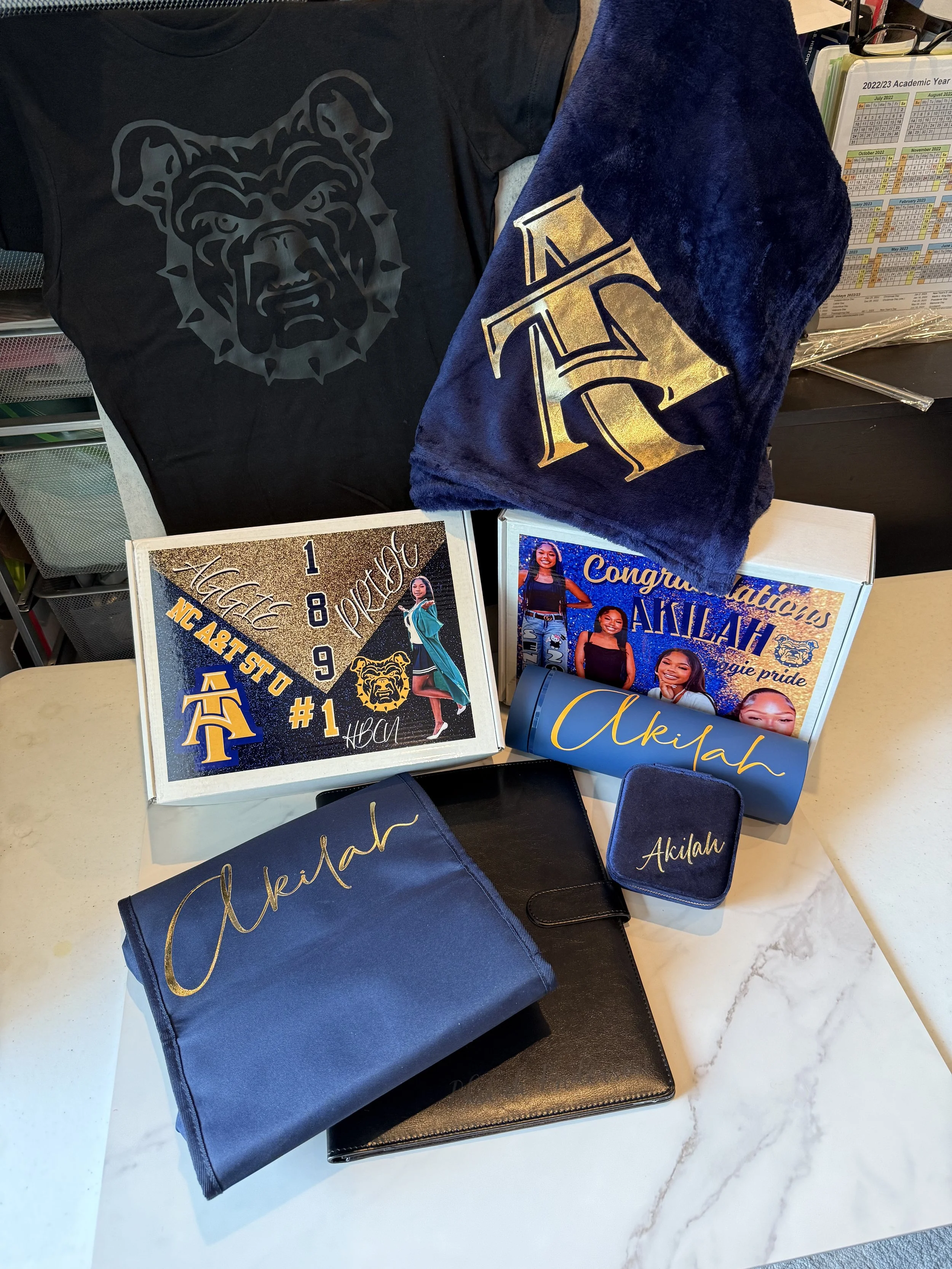 A collection of items celebrating a graduation, including a black shirt with a bulldog logo, a blue towel with a gold letter 'A', a personalized box with photos and the name 'Akhah', a blue pen holder with 'Akhah', a black planner, and a blue cloth w