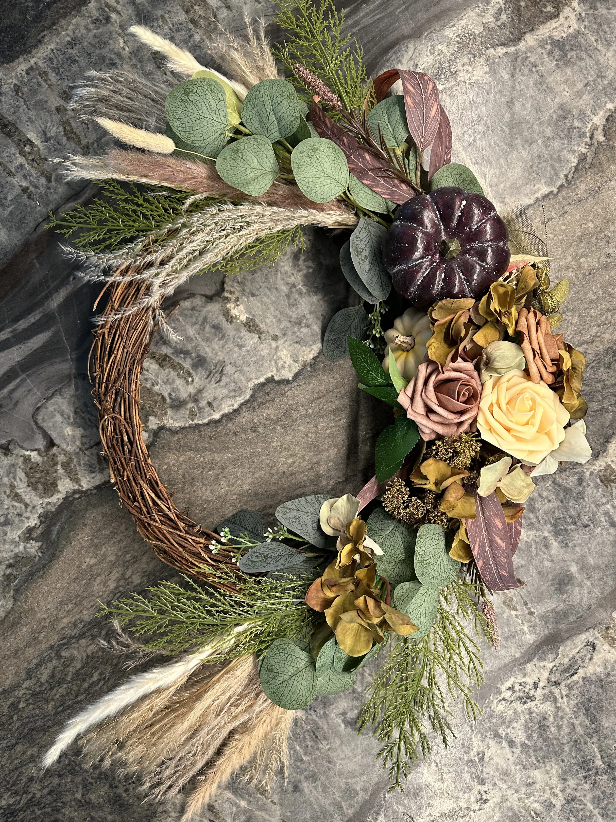 A decorative wreath made of woven branches, decorated with faux flowers, greenery, and a small dark purple pumpkin, set against a stone background.