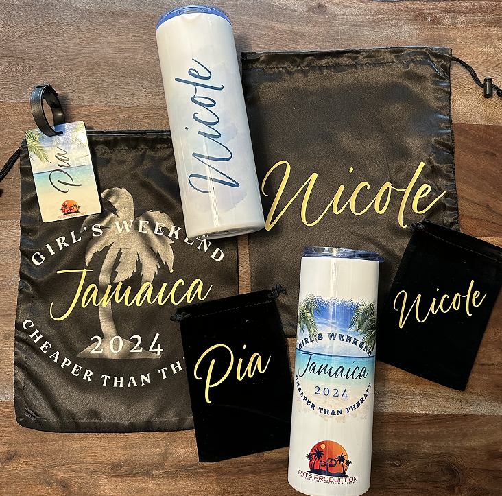 Items for Girl's Weekend in Jamaica 2024, including two white insulated bottles with tropical designs, a black drawstring bag with yellow and white text, and two smaller black pouches on a wooden surface.