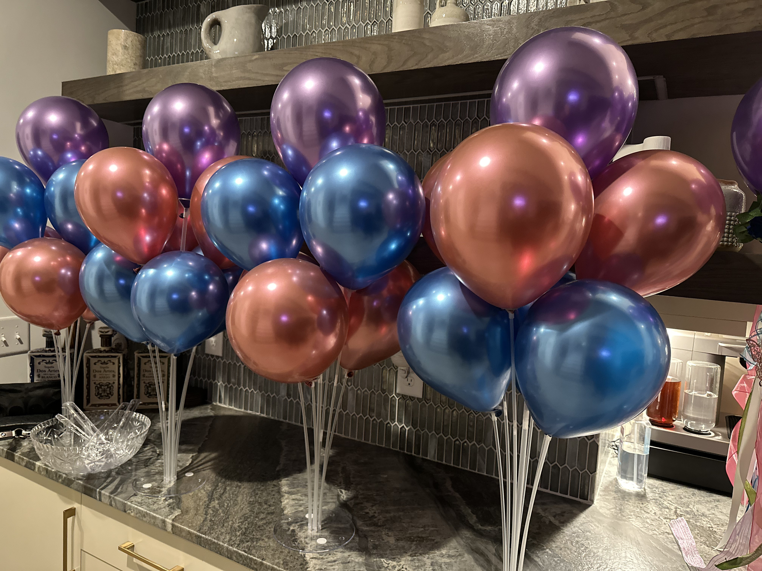 A bunch of metallic balloons in rose gold, purple, and blue colors, arranged on a kitchen countertop for decoration or celebration.
