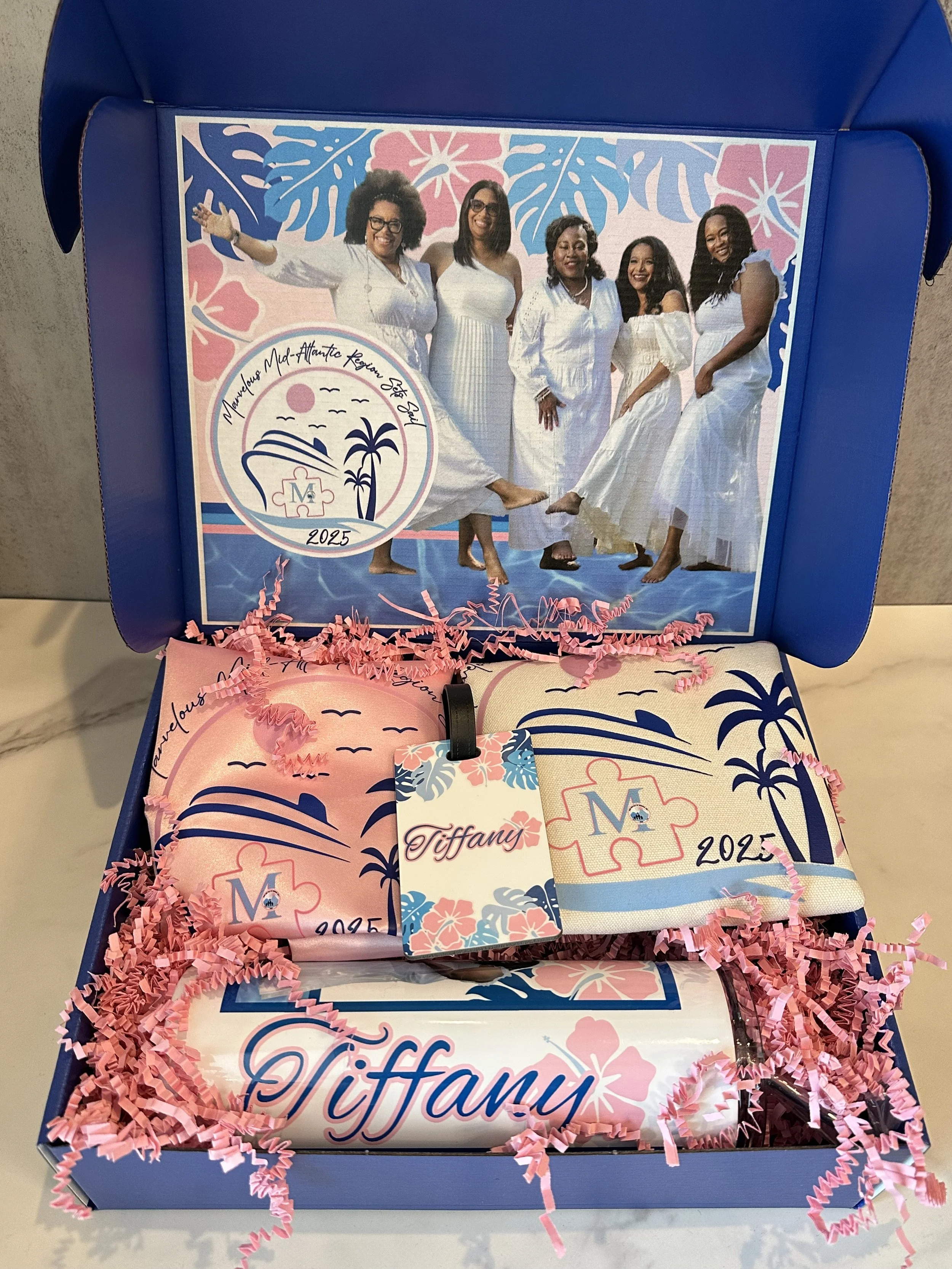 Open box with themed items for Tiffany, featuring a picture of six women in white dresses and a logo with a sun, palm trees, and ocean, and pink and blue decorations for a 2025 event.
