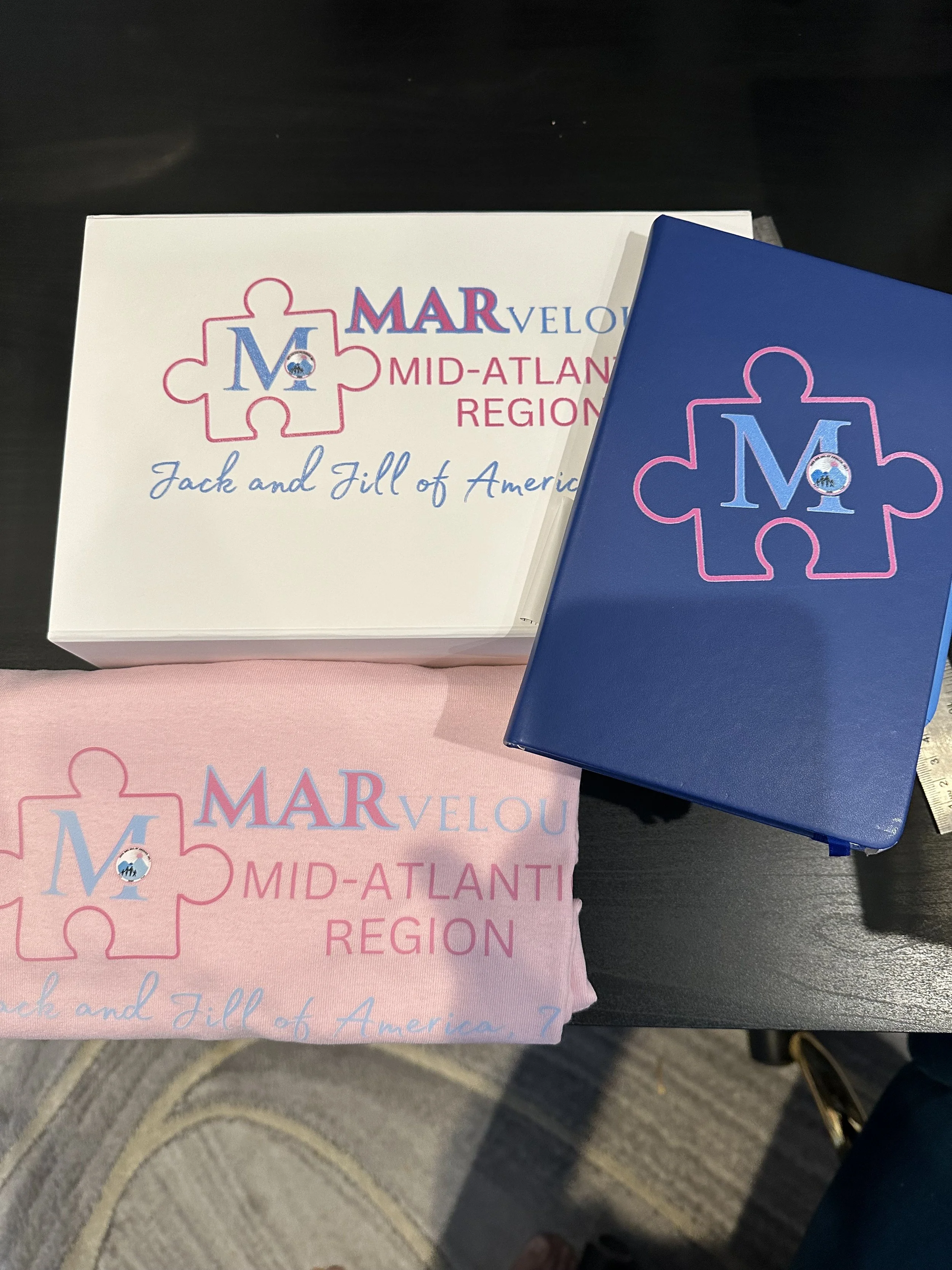 Collection of items related to the Mid-Atlantic Region of the Mar Velou organization, including a pink cloth, a white box, and a blue box, all featuring the organization’s logo with a puzzle piece and a globe, and the text 'Jack and Jill of America'.