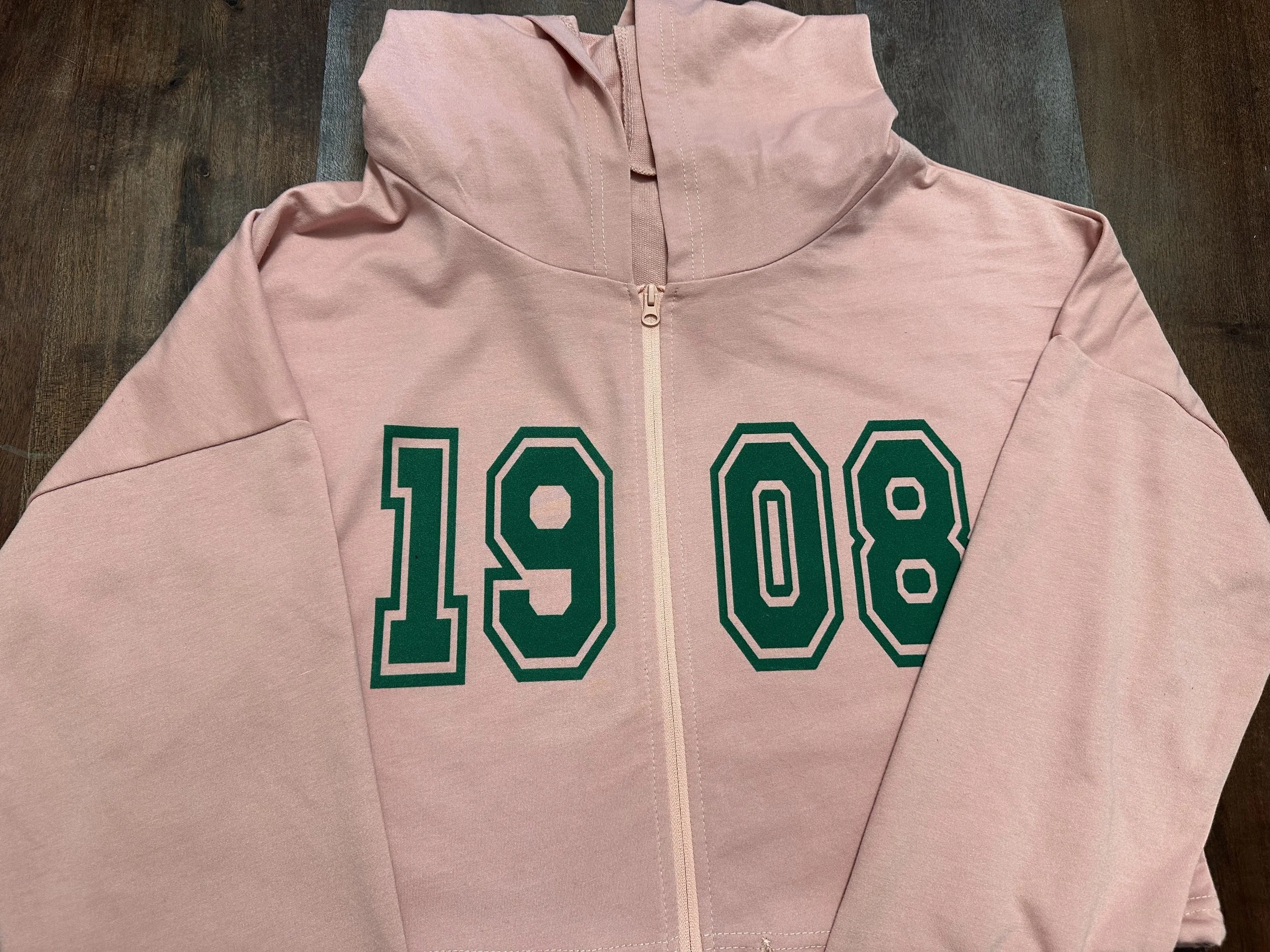 A pink hoodie with a front zipper, featuring the numbers '1908' printed in large green outlined font on the chest, laying on a wooden surface with a gray mat underneath.