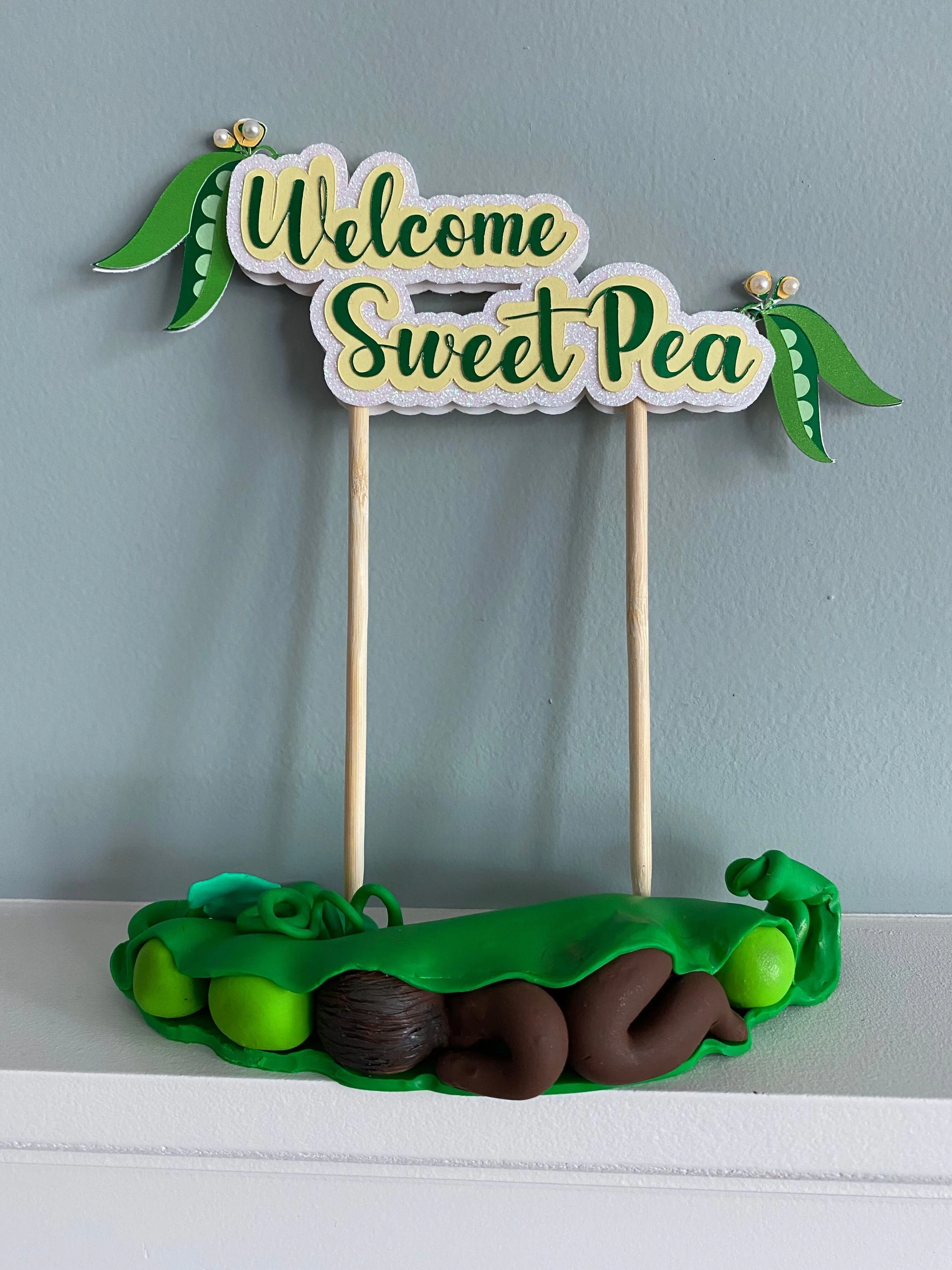 Decorative sign that reads 'Welcome Sweet Pea' with green leaves and yellow flowers, attached to wooden sticks, sitting on a crocheted green blanket with green and brown yarn balls around it.