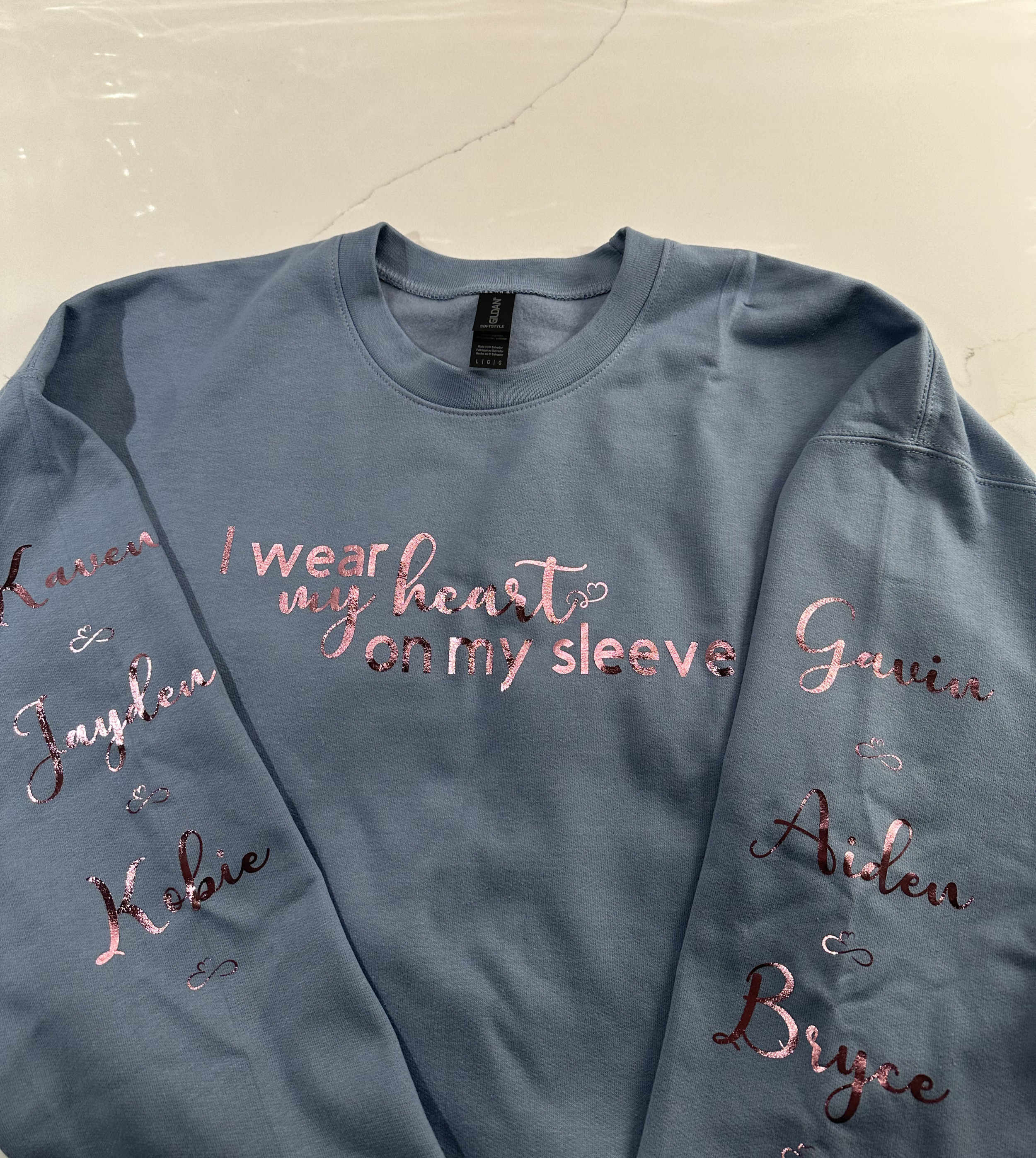 A light blue sweatshirt with pink embroidered text that reads 'I wear my heart on my sleeve' in the center, and names embroidered on the sleeves including 'Karen,' 'Jaylen,' 'Kobie,' 'Gavin,' 'Aiden,' and 'Bruce,' each separated by small decorative hearts.