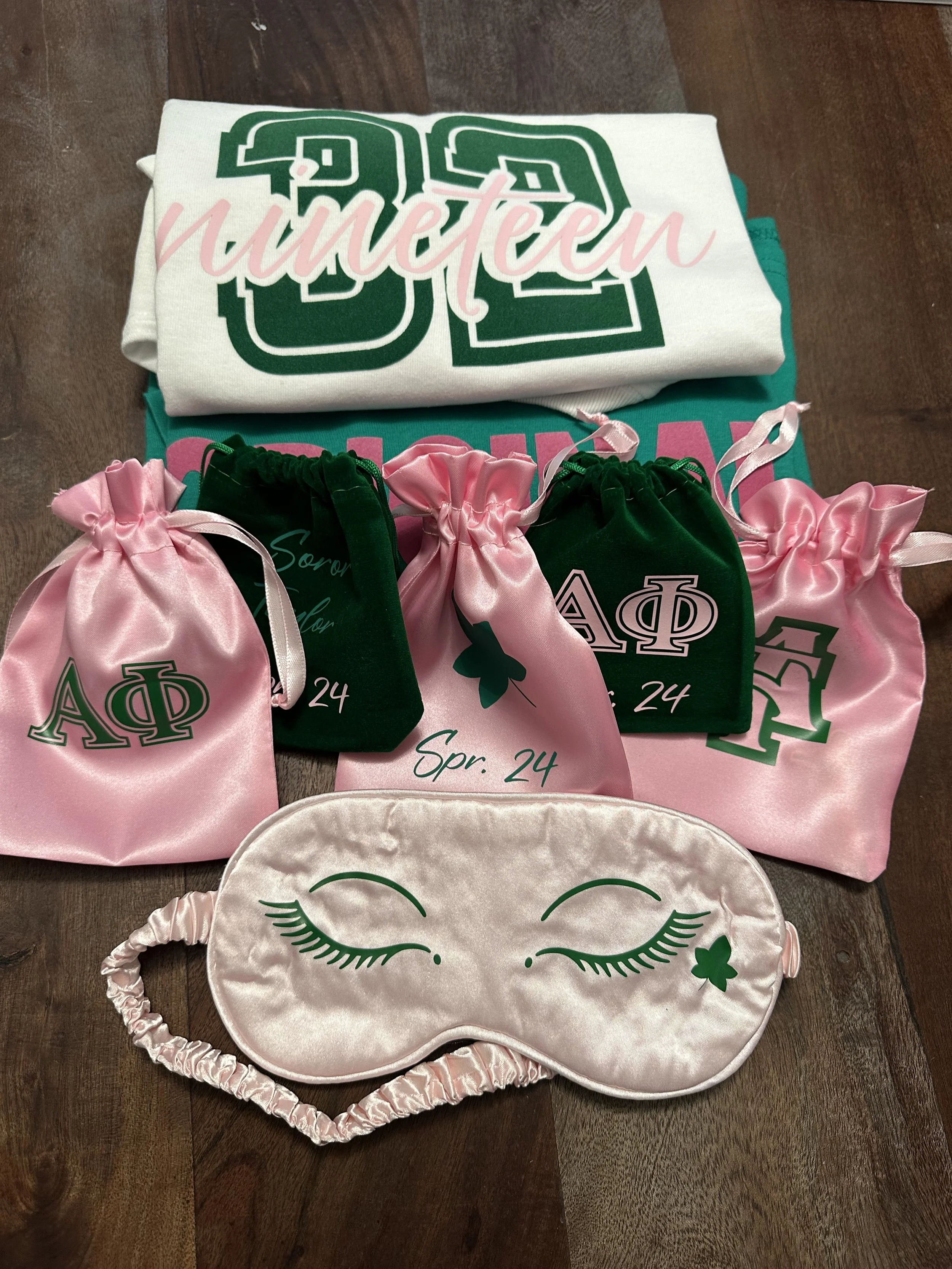 A collection of sorority memorabilia including a white t-shirt with green and pink text, pink and green satin pouches with Greek letters and the date 'Apr. 24', and a pink sleep mask with closed eyes and eyelashes design, all laid out on a wooden sur
