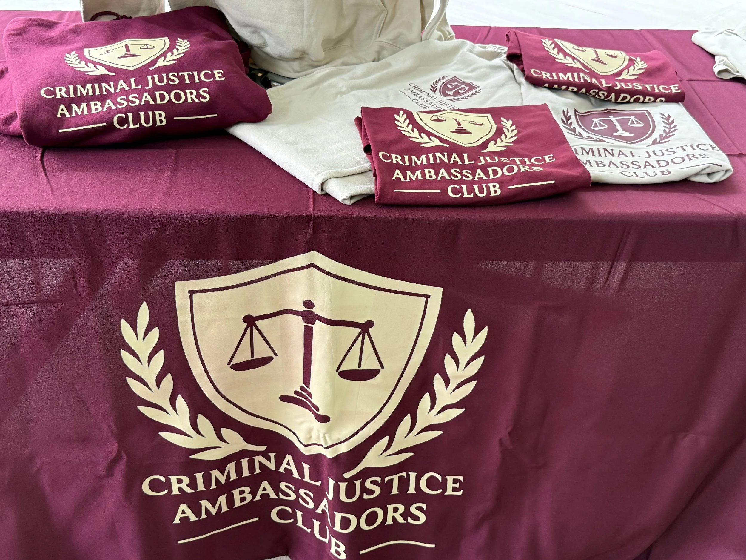 A table covered with a maroon tablecloth featuring the logo of the Criminal Justice Ambassadors Club. On the table, there are several folded maroon and white T-shirts with the club's emblem and name printed on them.