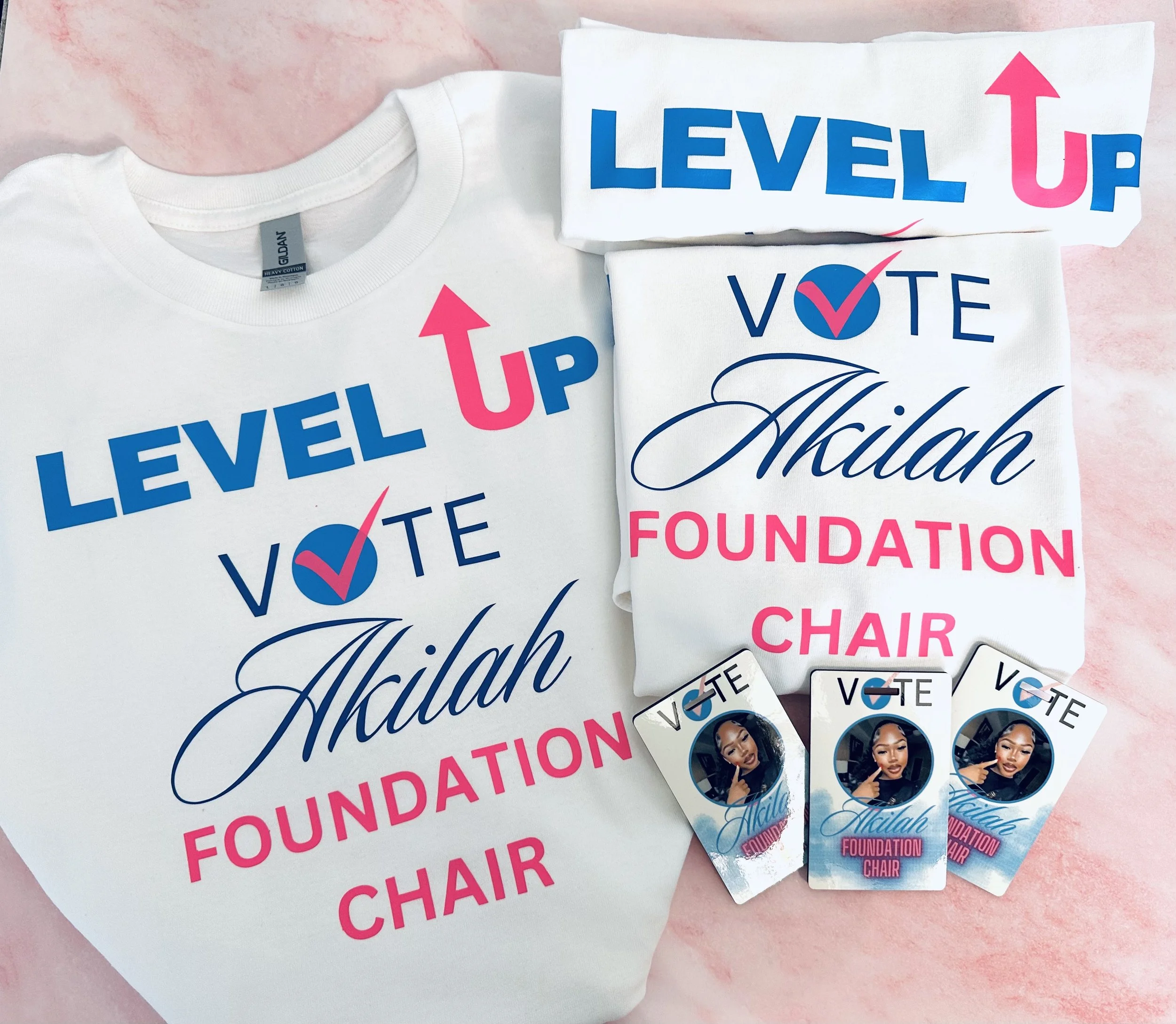 Campaign merchandise for Akilah Foundation, including white t-shirts with blue and pink text, campaign buttons featuring a woman’s photo, and white tote bags with the same design.