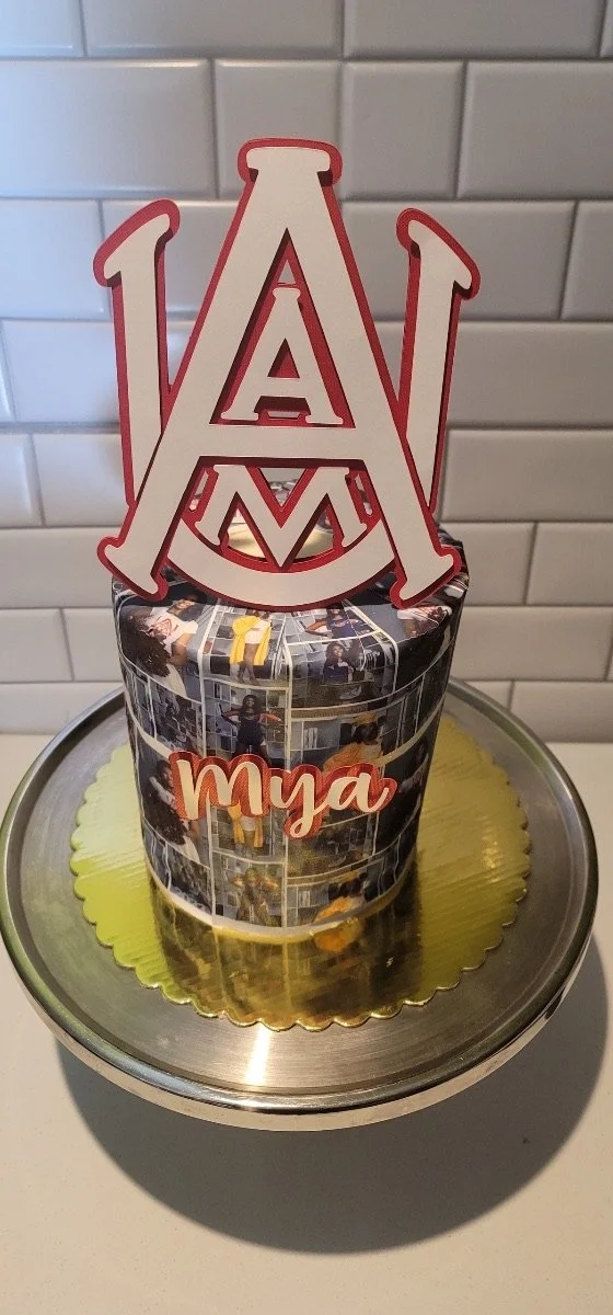 A cake with a collage of photos and a large red and white decoration on top, featuring a prominent letter A, with the name 'Mya' written in red on the front. The cake is placed on a gold-colored board and is on a silver scale or cake turntable, with 