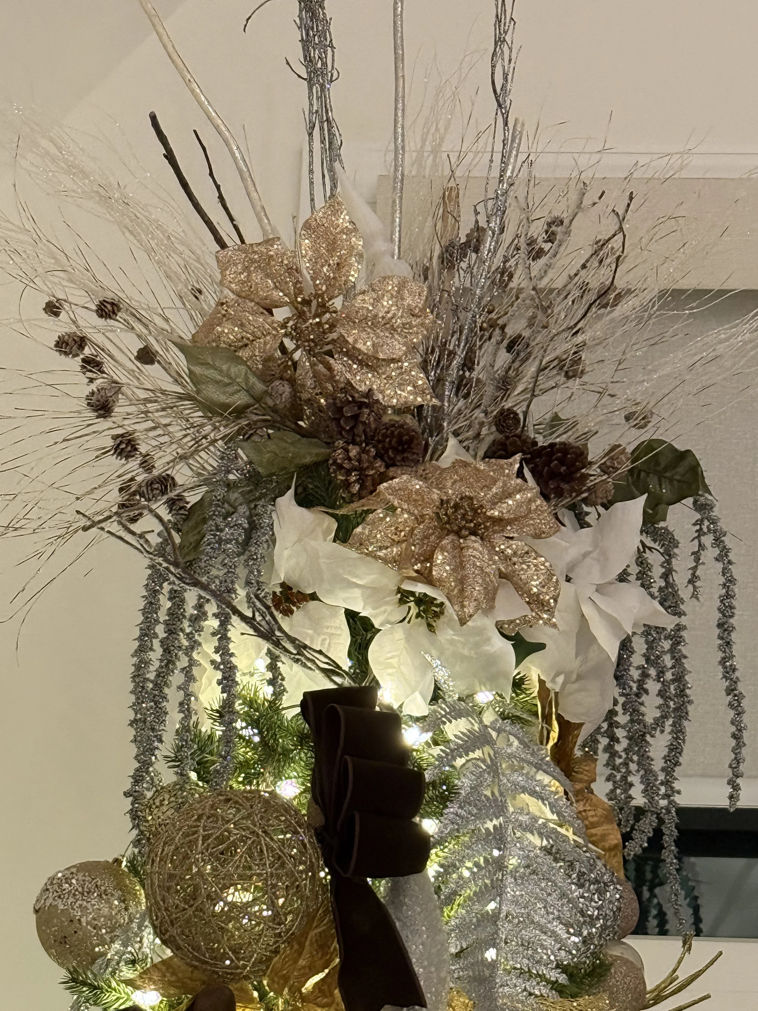 Decorative Christmas arrangement with gold and silver ornaments, white poinsettias, gold poinsettias, pinecones, greenery, and lights.