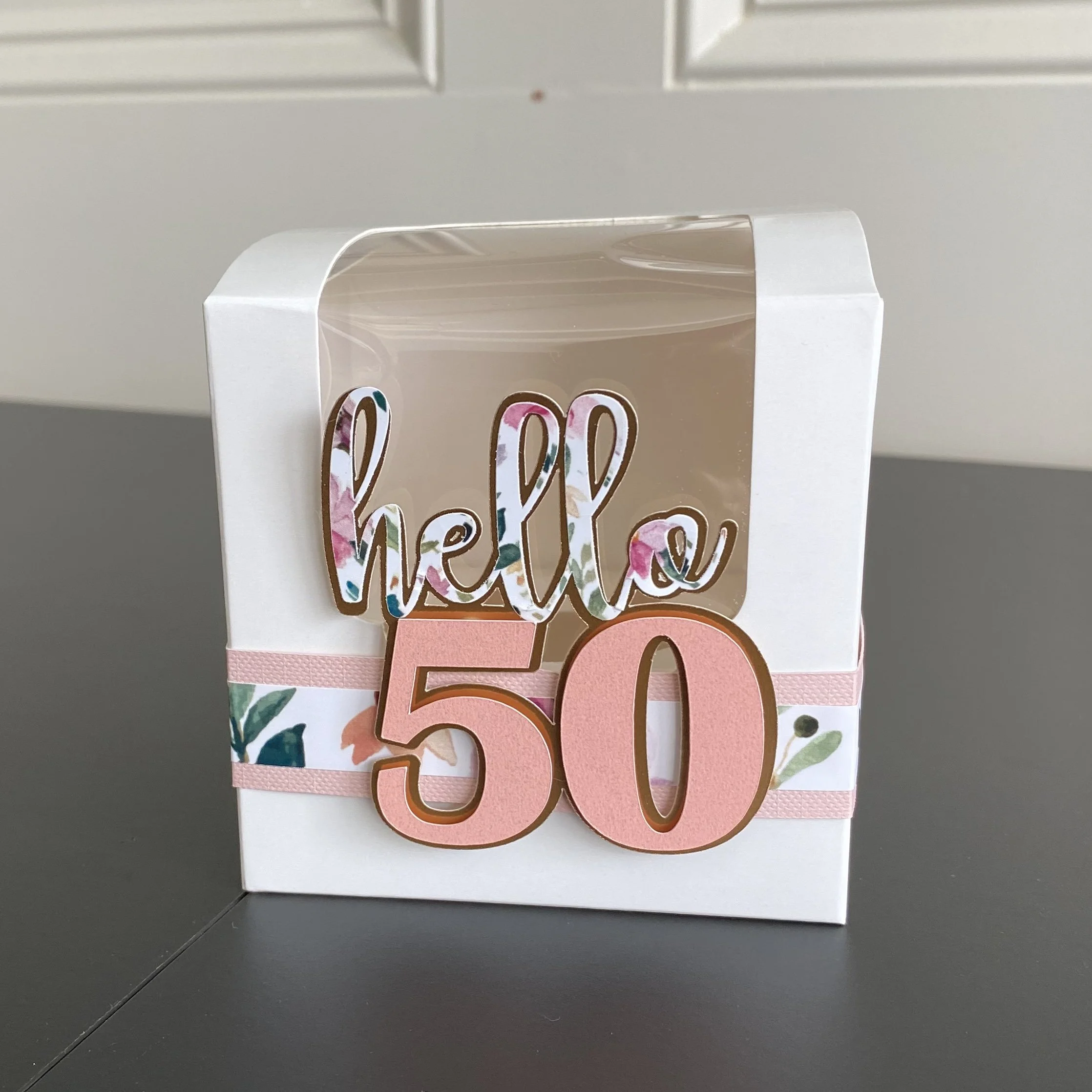 Decorative cake topper with the words 'hello' in floral design and '50' in pink, mounted on a white and floral decorated box.