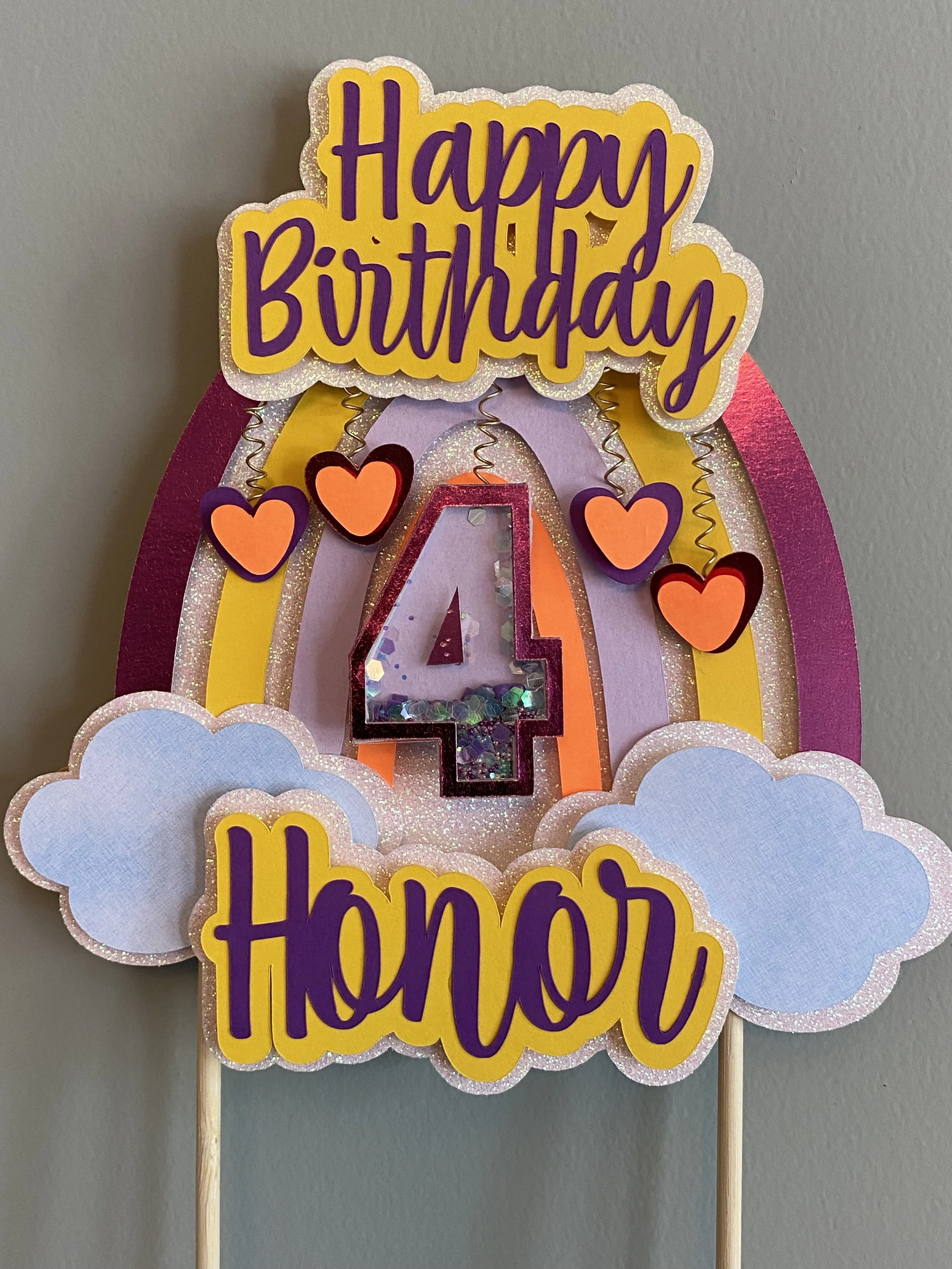 A colorful birthday decoration with a rainbow, pink, yellow, and purple colors, featuring signs that say "Happy Birthday" and "honor," a large glittery number "4", and clouds and hearts.