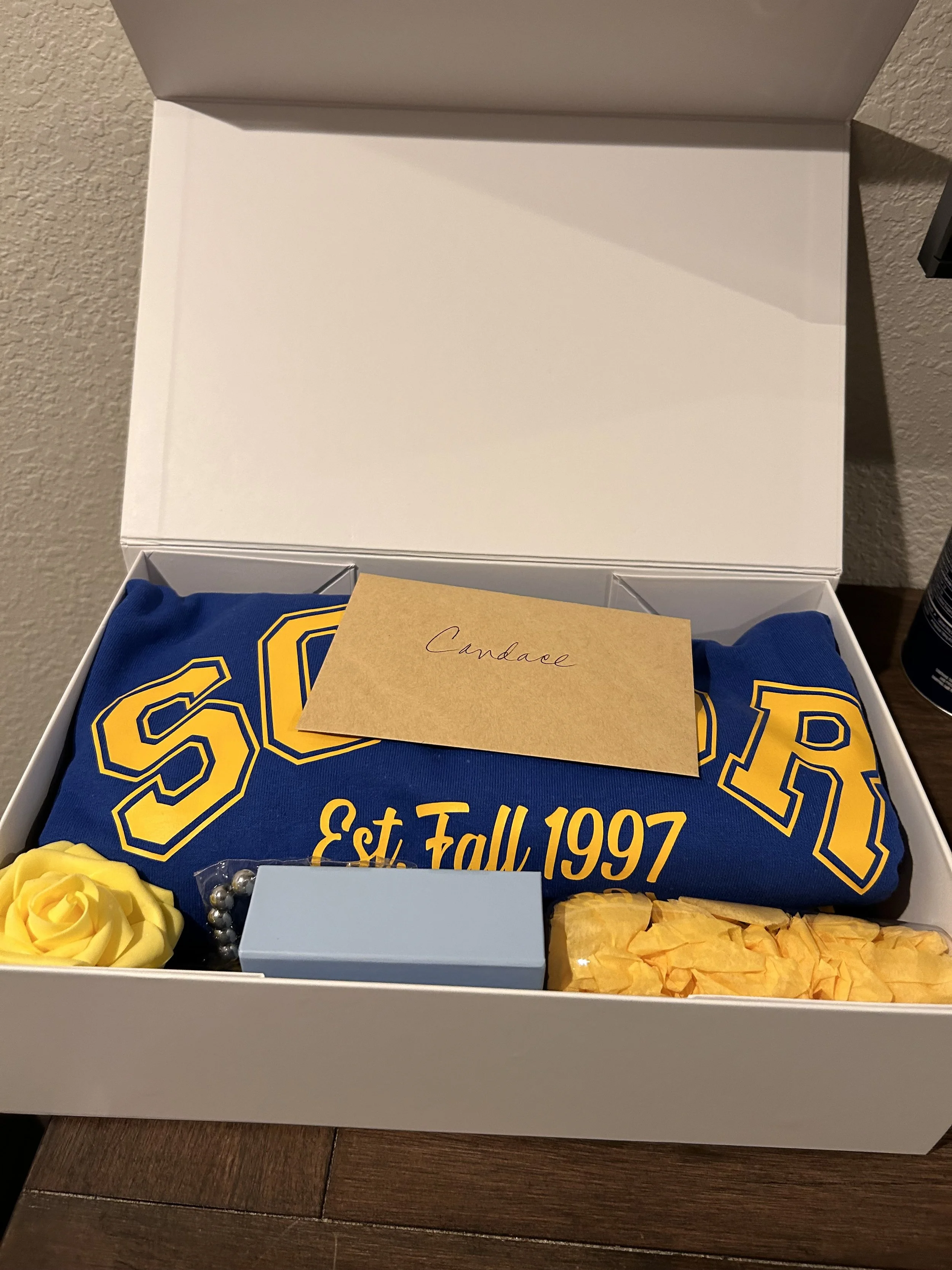 A white box with a blue sweatshirt that has yellow lettering reading "S O R R" and "Est. Fall 1997" inside. The box contains a yellow artificial rose, a gray jewelry box, a silver beaded necklace, and yellow tissue paper. A brown note with handwritte