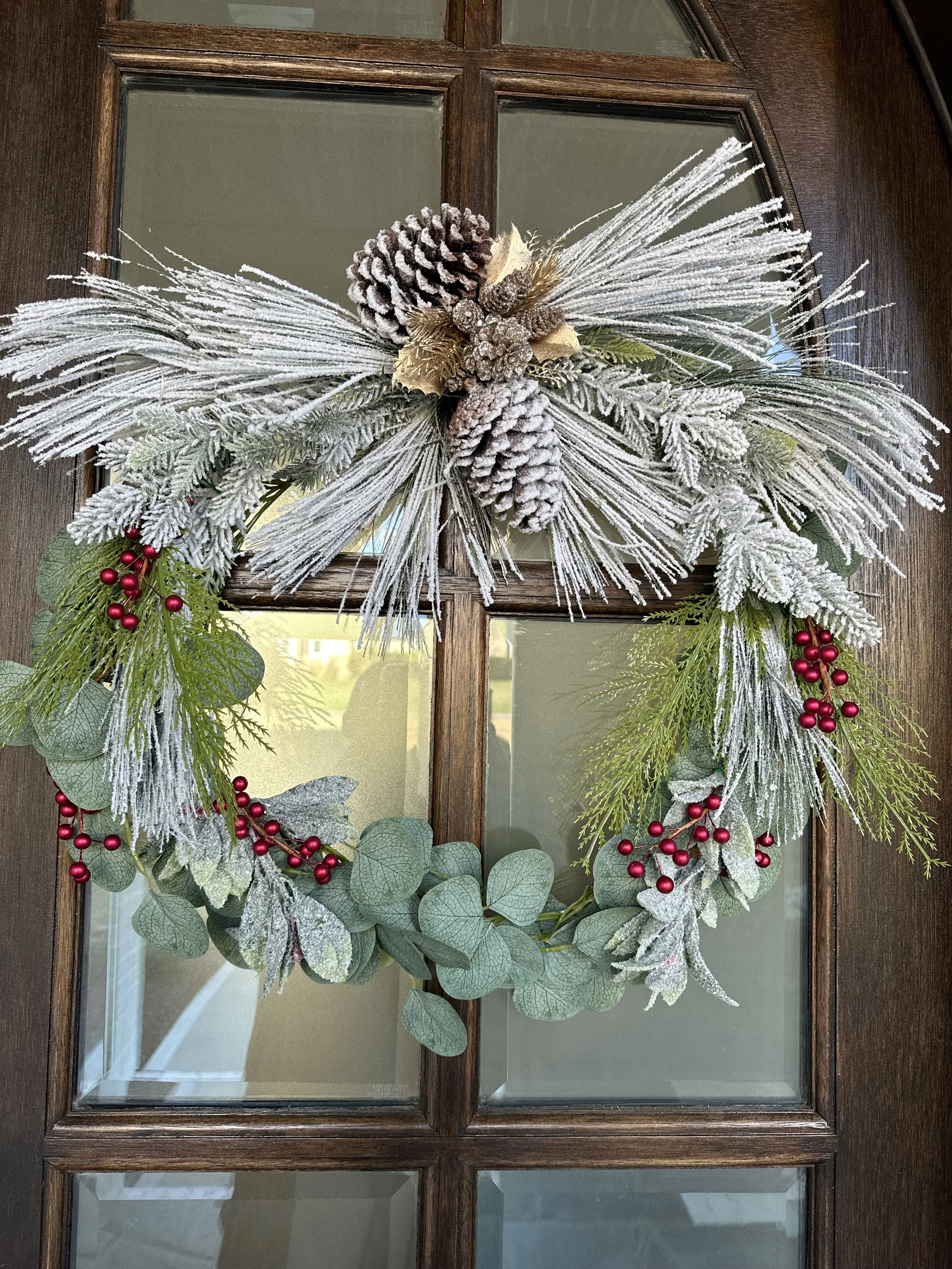 Holiday wreath decorated with snow-dusted pine cones, frosted greenery, red berries, and gold accents hanging on a wooden door.