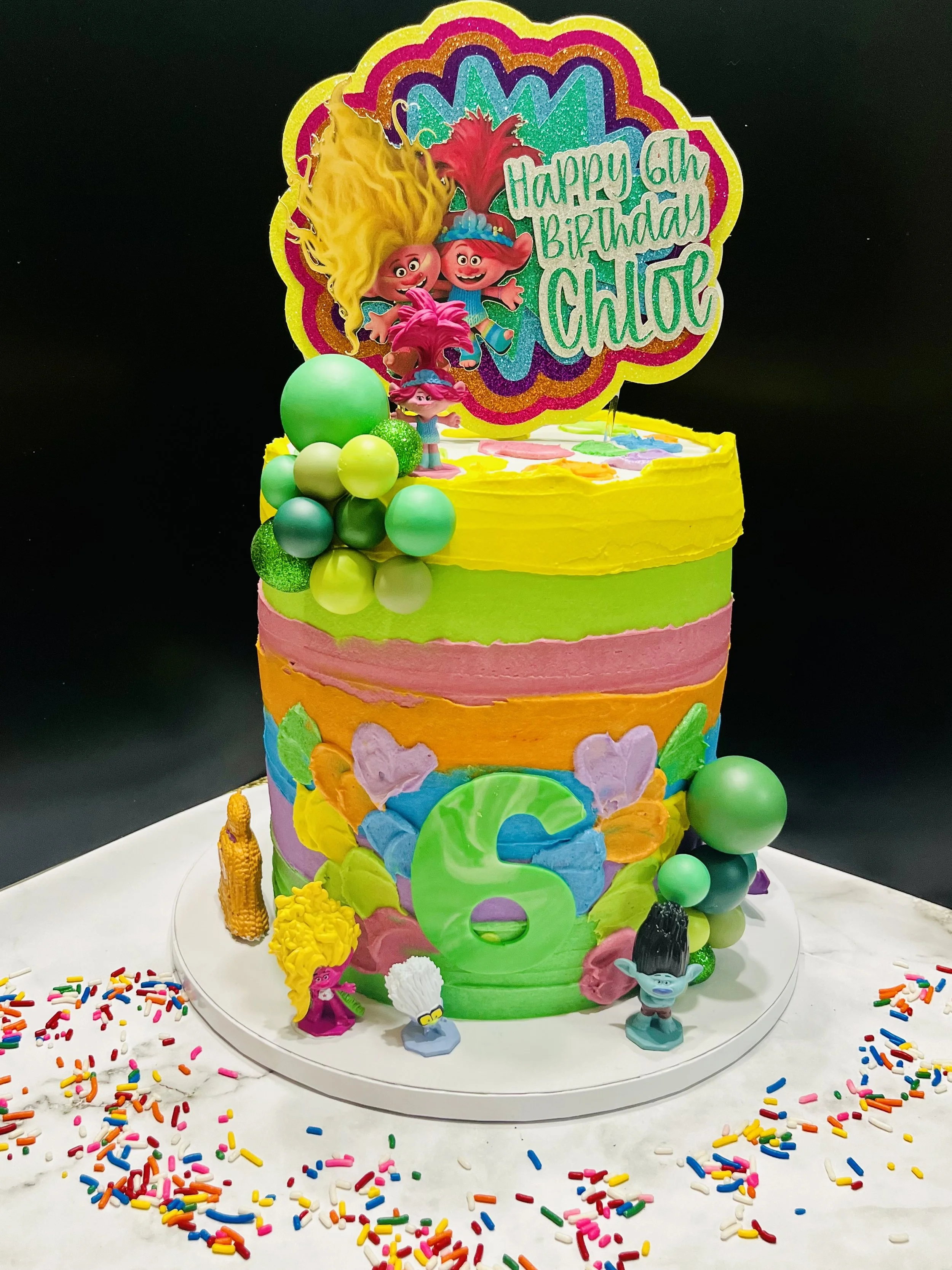 Colorful birthday cake decorated with Trolls characters, balloons, and sprinkles. It has a large topper with a message for Chloe's 6th birthday.