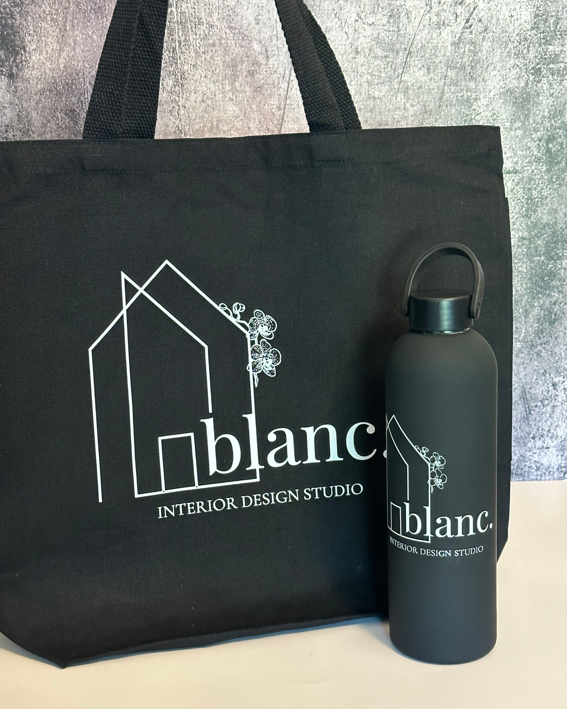 A black tote bag and a matching black water bottle both with the logo and text of 'blanc. INTERIOR DESIGN STUDIO' on them, placed on a surface with a textured background.