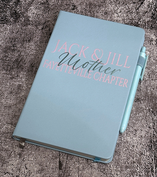 Light blue notebook with gold and pink lettering and a matching pen attached, resting on a textured gray surface.