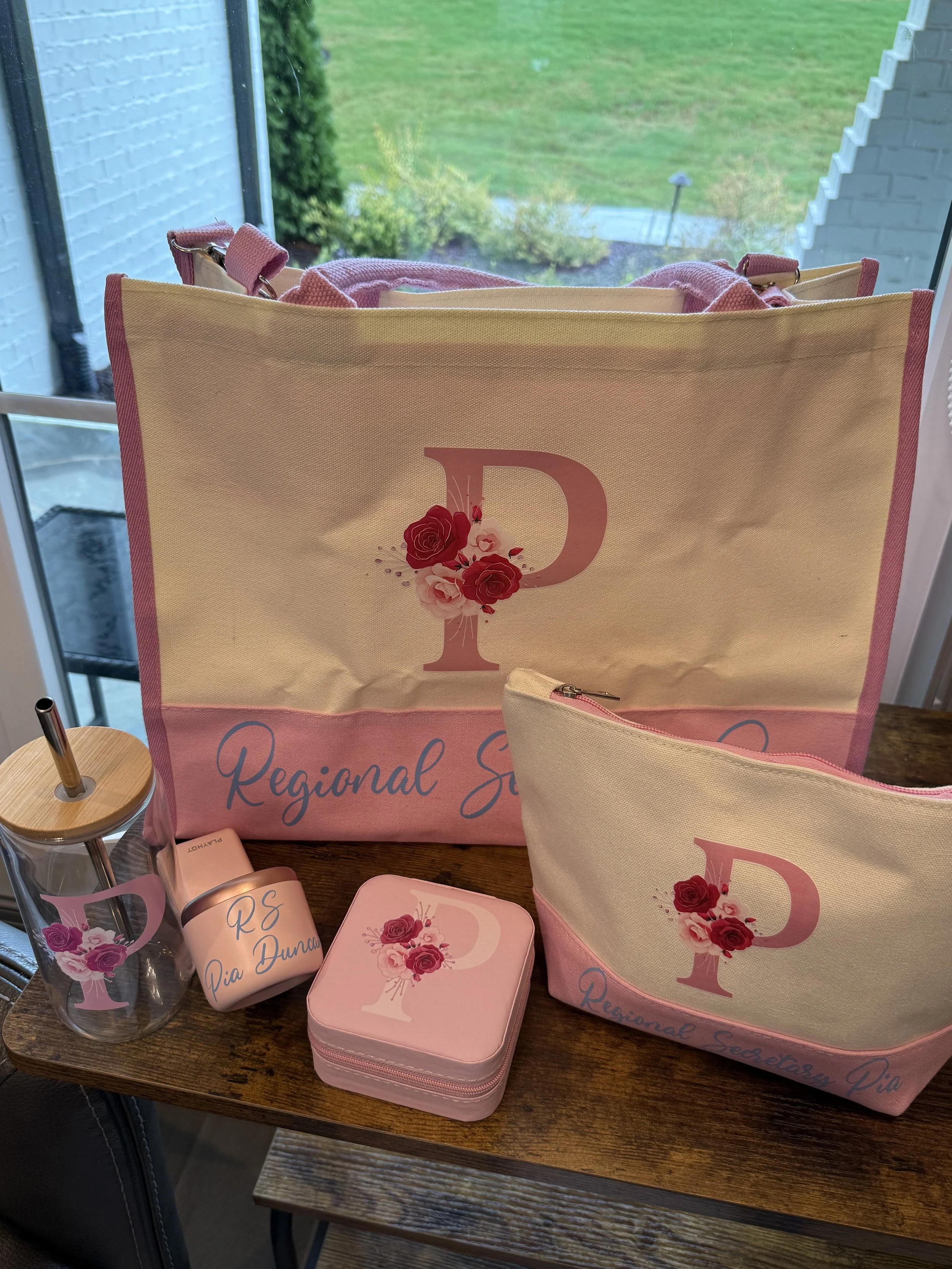 Collection of pink and cream tote bag, small pouch, water bottle with pink floral decal, and pink enclosed container on a wooden table. The tote bag and pouch have a large letter 'P' with pink roses and the text 'Regional Secretary Pia' visible on th