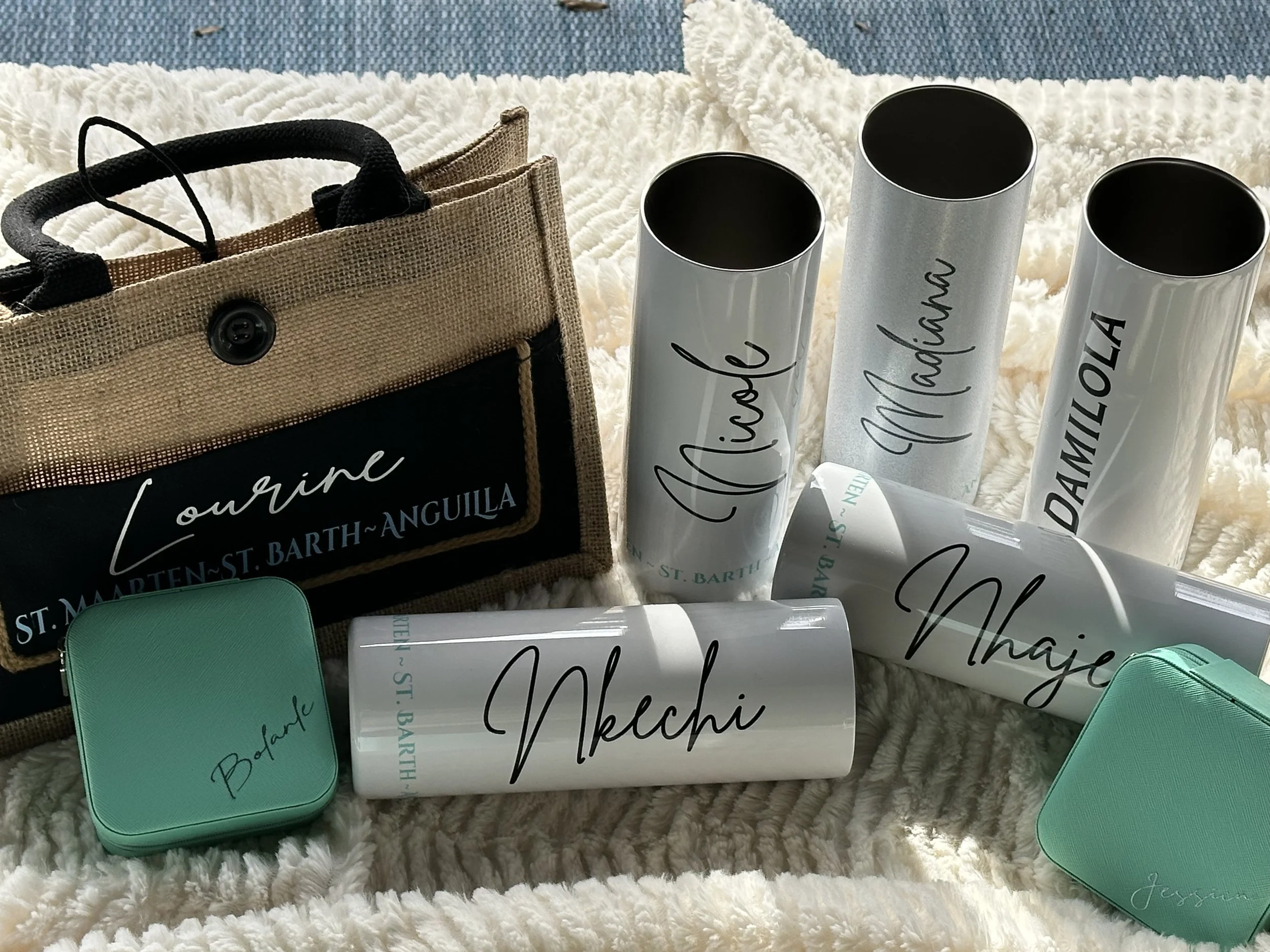 Travel souvenir collection on a soft white blanket, including a tan tote bag with black lettering, two mint green tins, and five white tumblers with black cursive names of different islands, such as 'Michele', 'Martinique', 'Damilola', 'Nkechi', and 
